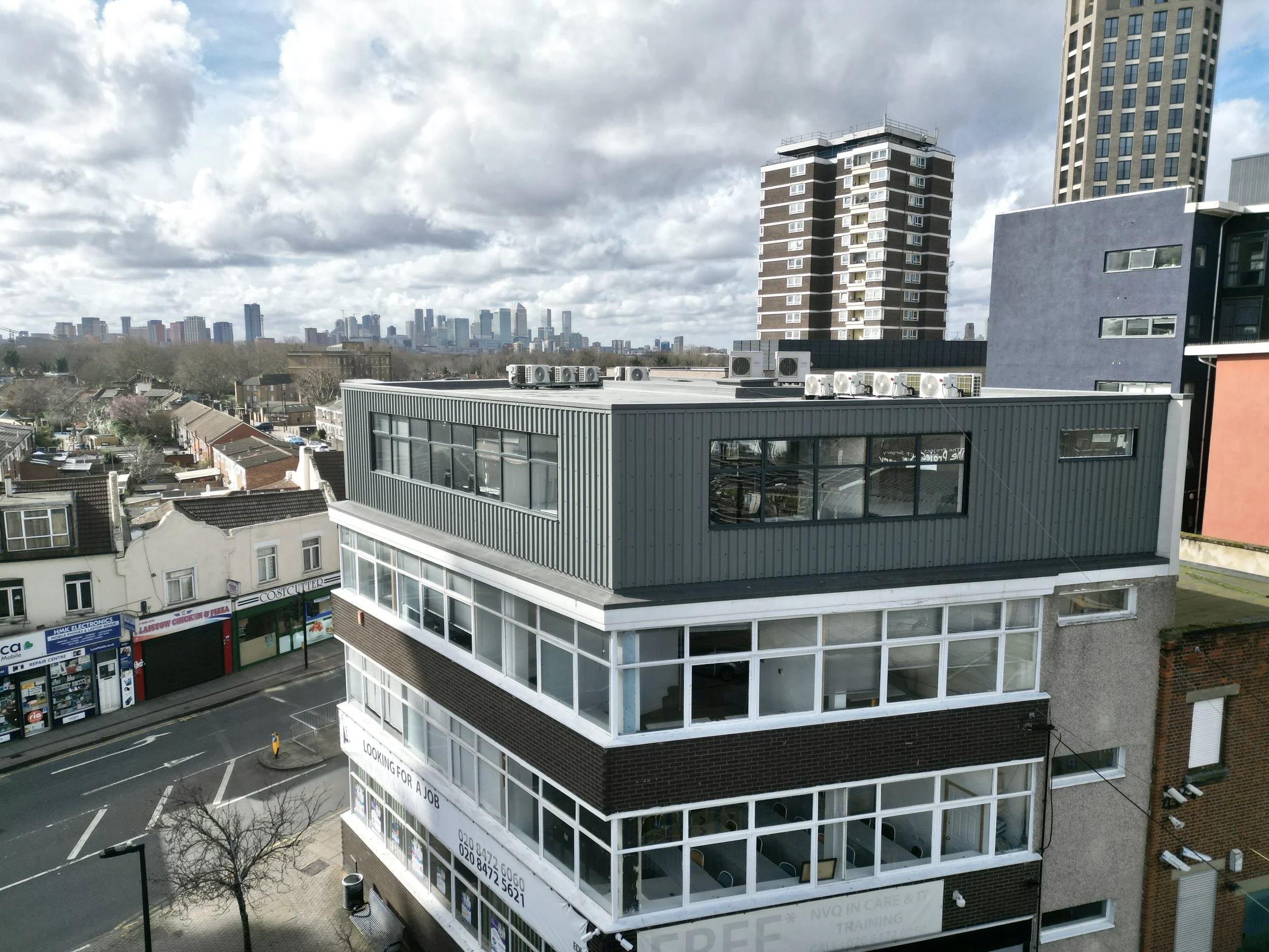 View of urban buildings with a mix of residential and commercial structures, foreground with a multi-story building with large windows, rooftop air conditioning units, street lined with businesses and stores, distant city skyline, cloudy sky.