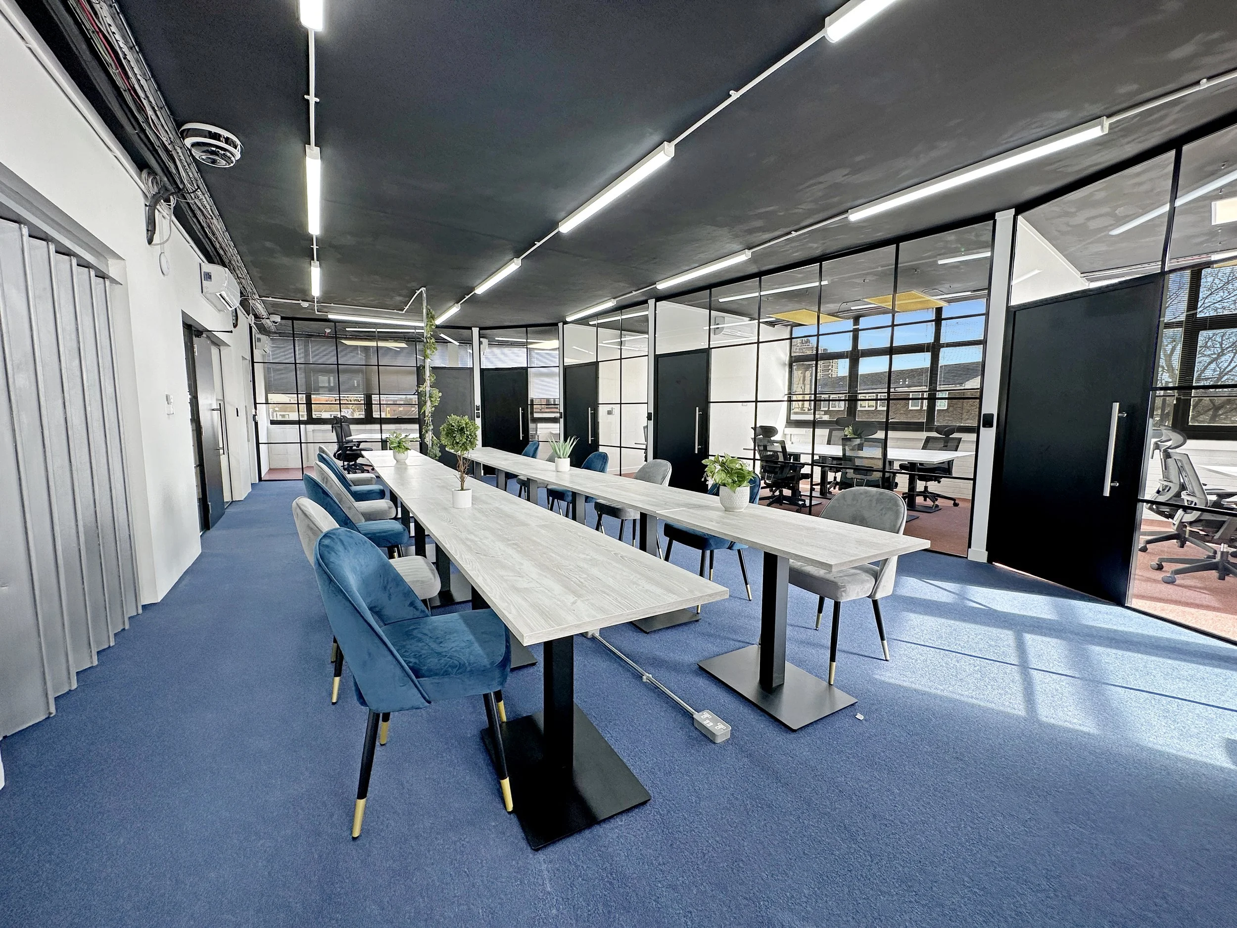 Modern office space with long tables and chairs, glass partition walls, blue carpet, and ceiling lights.