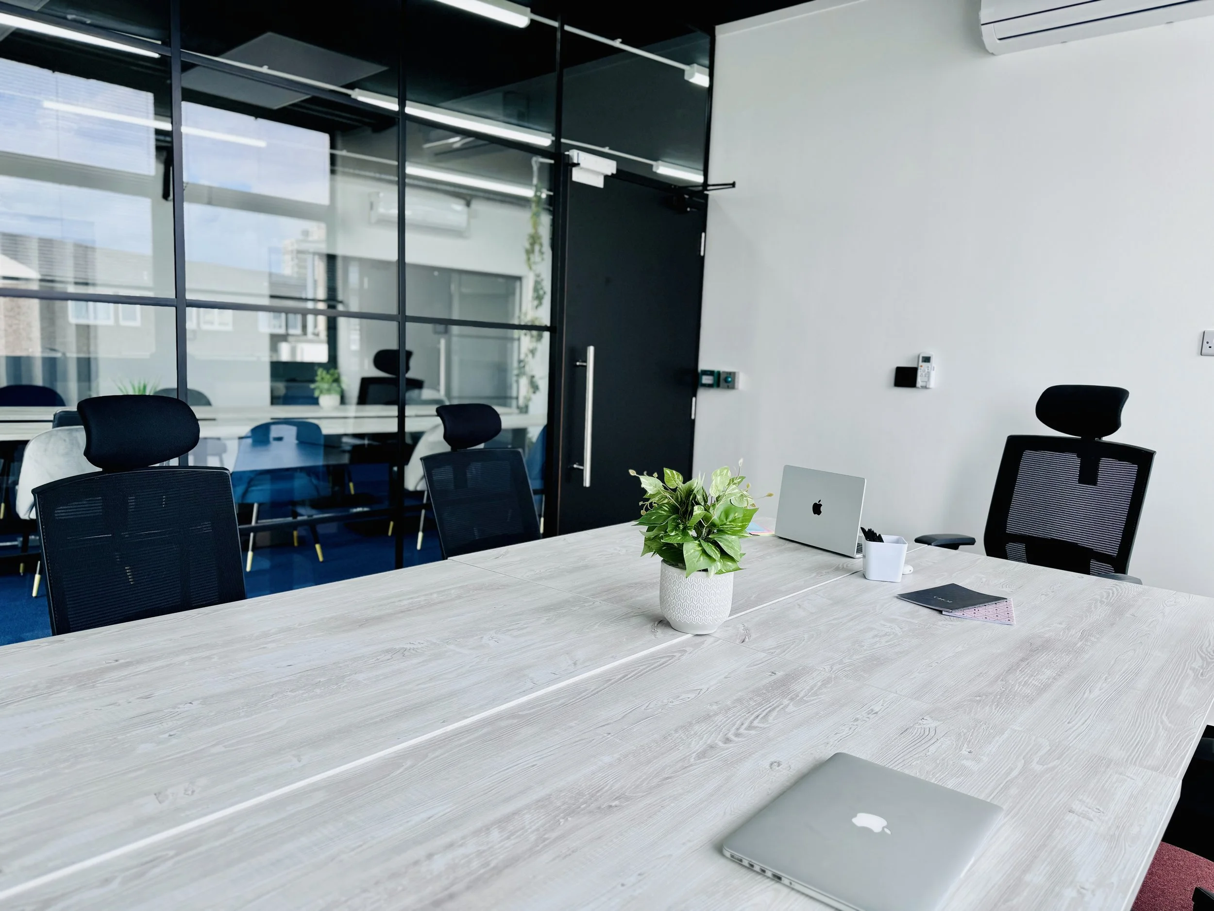 Modern office meeting room with a large wooden table, black mesh chairs, a laptop, a potted plant, and a glass partition.