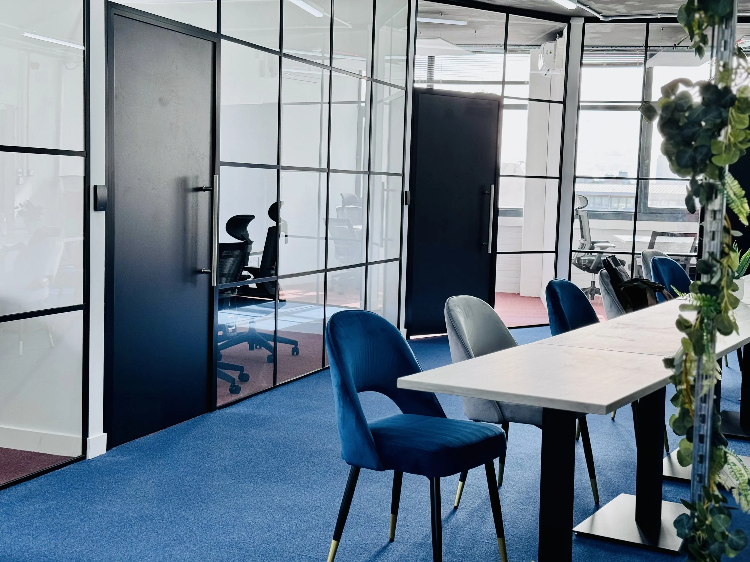 Modern office space with glass partitions, blue carpet, conference table, blue and gray chairs, and plants. Black office chairs visible through partitions.