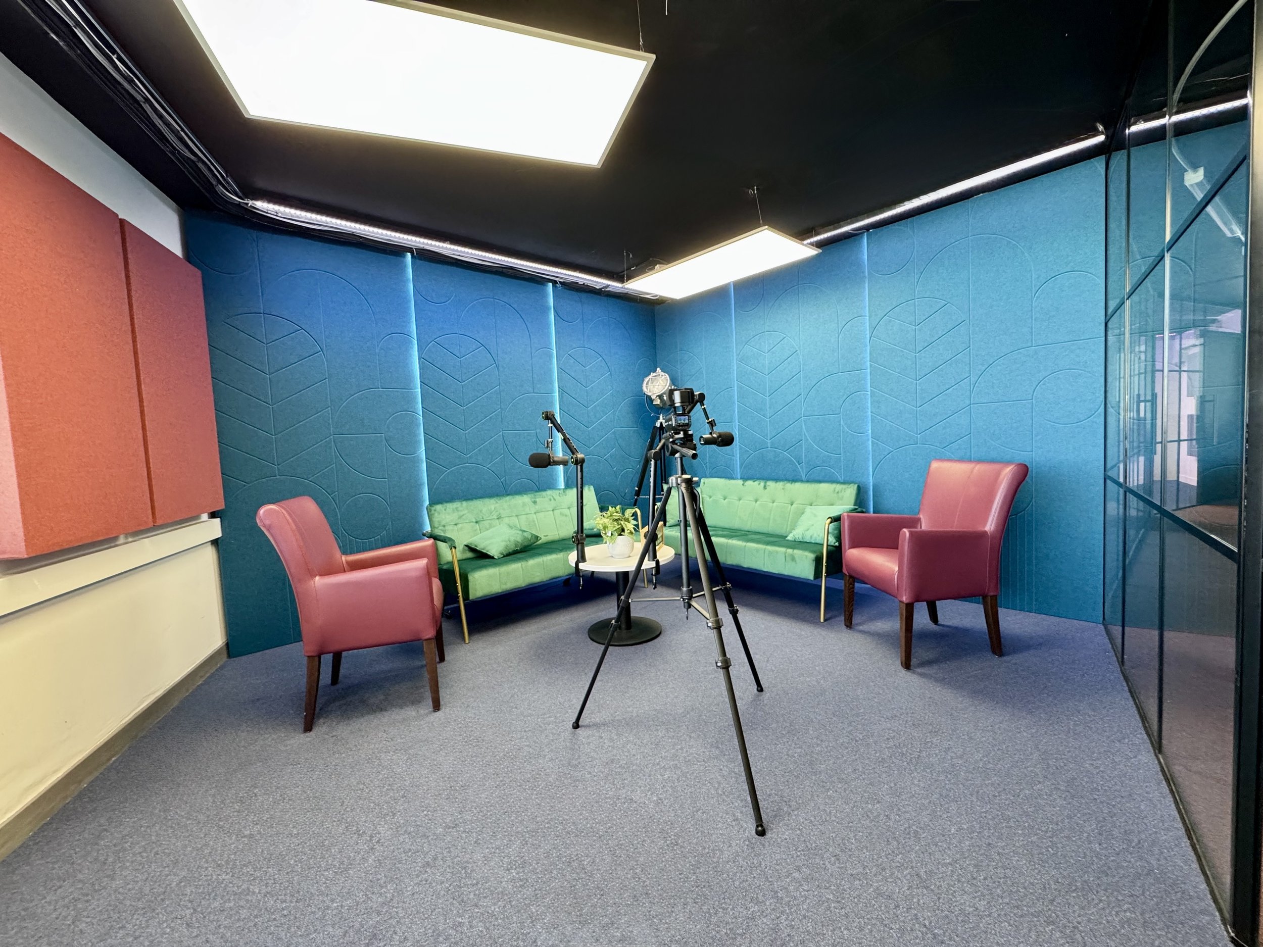 Podcast studio with pink chairs, green sofa, microphones, camera on tripod, blue acoustic panels, and white lighting.