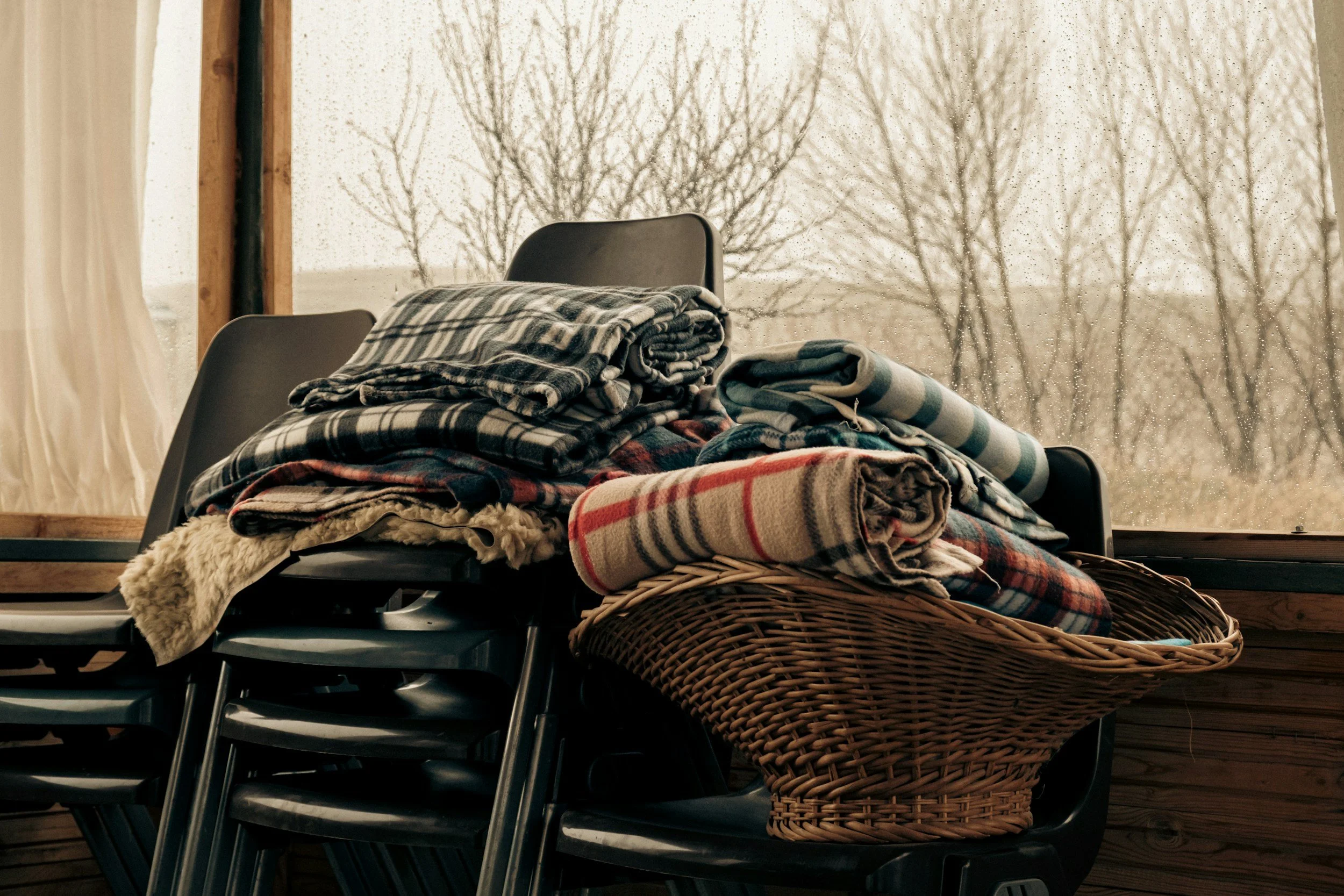 Stack of folded blankets and throws on chairs and in a wicker basket near a window with raindrops on the glass, looking outside at leafless trees.