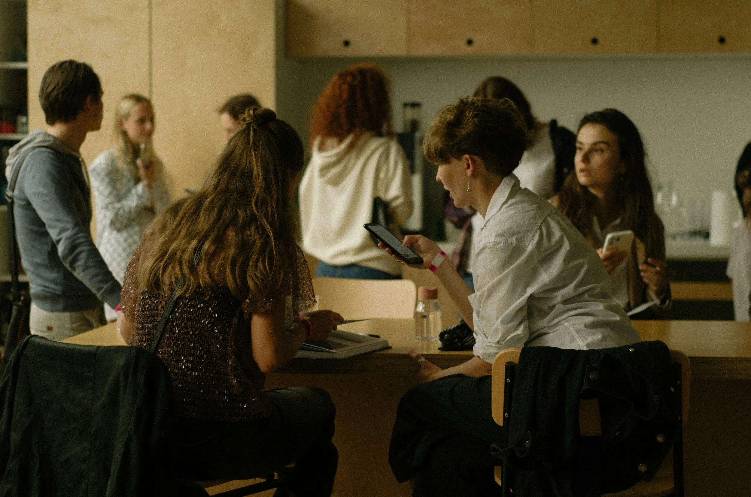 Group of people in a workshop some sitting at a table engaged in conversation and others standing in the background, some using smartphones.