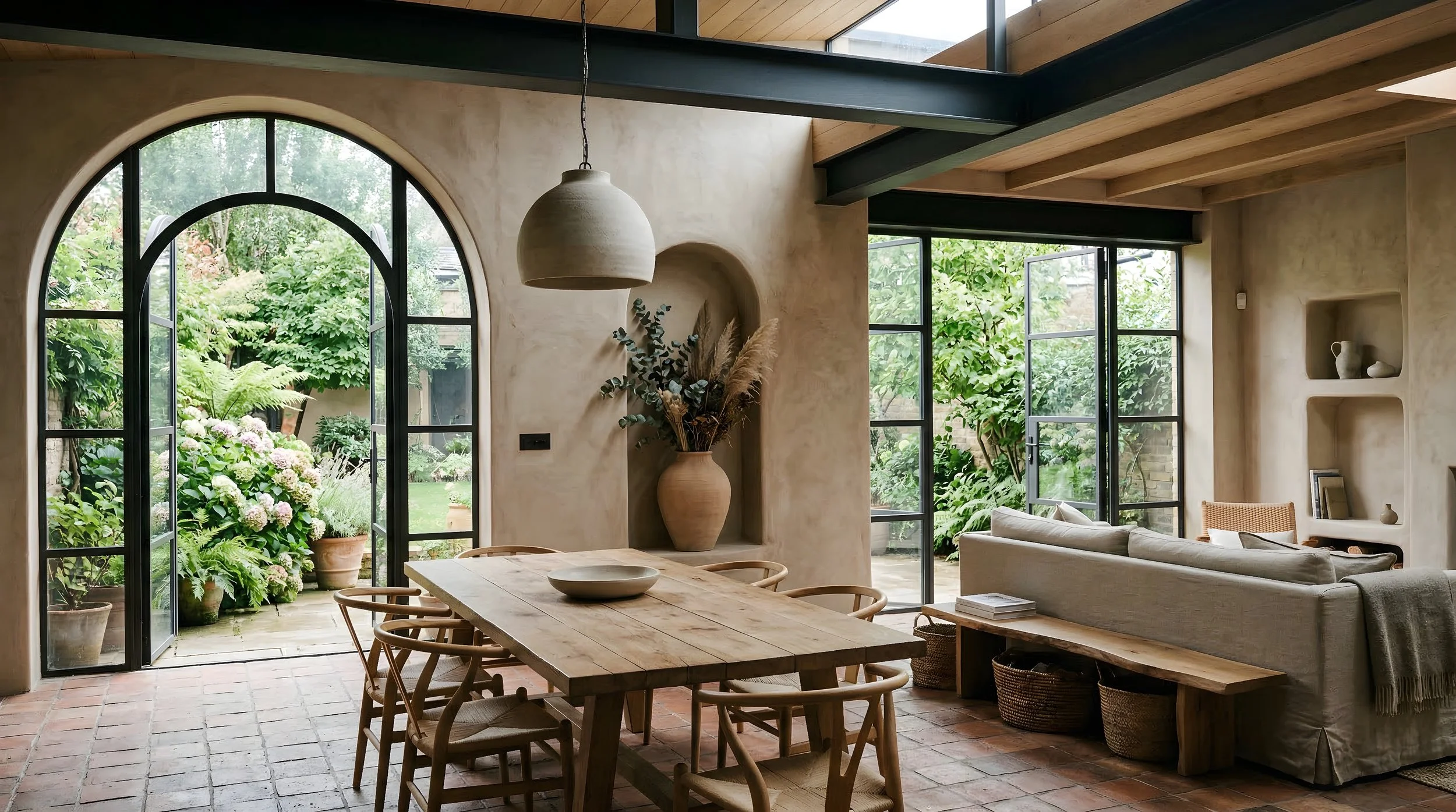 Open plan living and dining room with arched Crittall doors, lime plaster walls, terracotta tile floor and exposed steel beams.