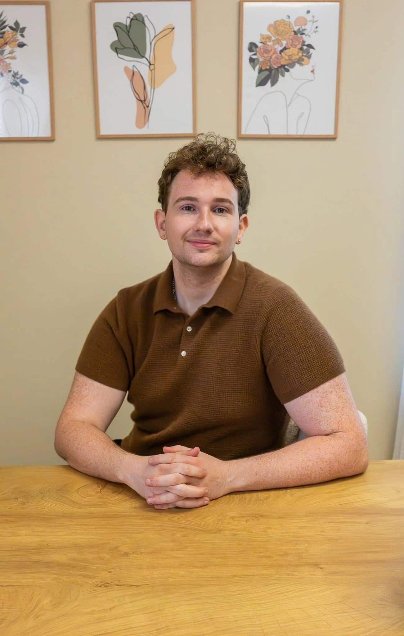 A person with curly hair sits at a wooden table, smiling, wearing a brown shirt. Three floral art pieces hang on the wall behind.