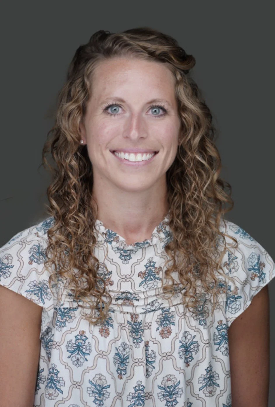 Woman with long curly hair and blue eyes, smiling, wearing a patterned short-sleeve top, on a plain gray background.
