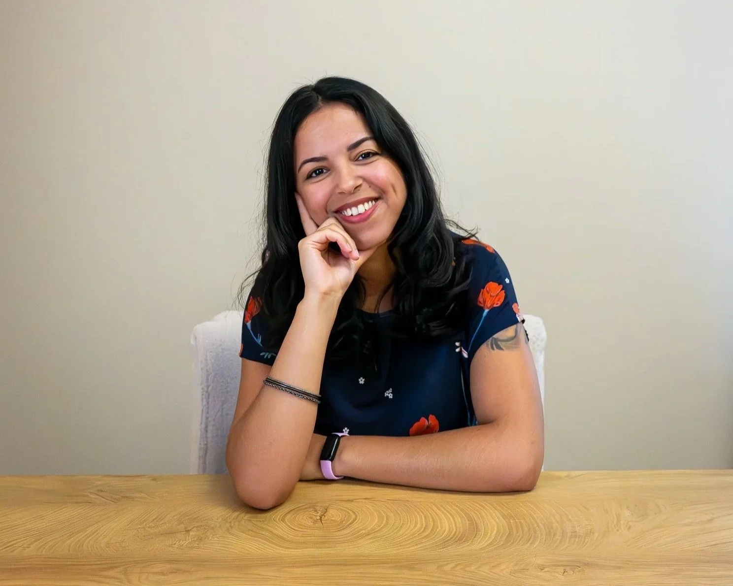Smiling woman with long dark hair sits at a wooden table, resting her chin on her hand, against a plain background.