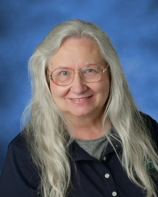 mature woman with glasses and long, wavy grey hair facing forward, smiling at the camera.