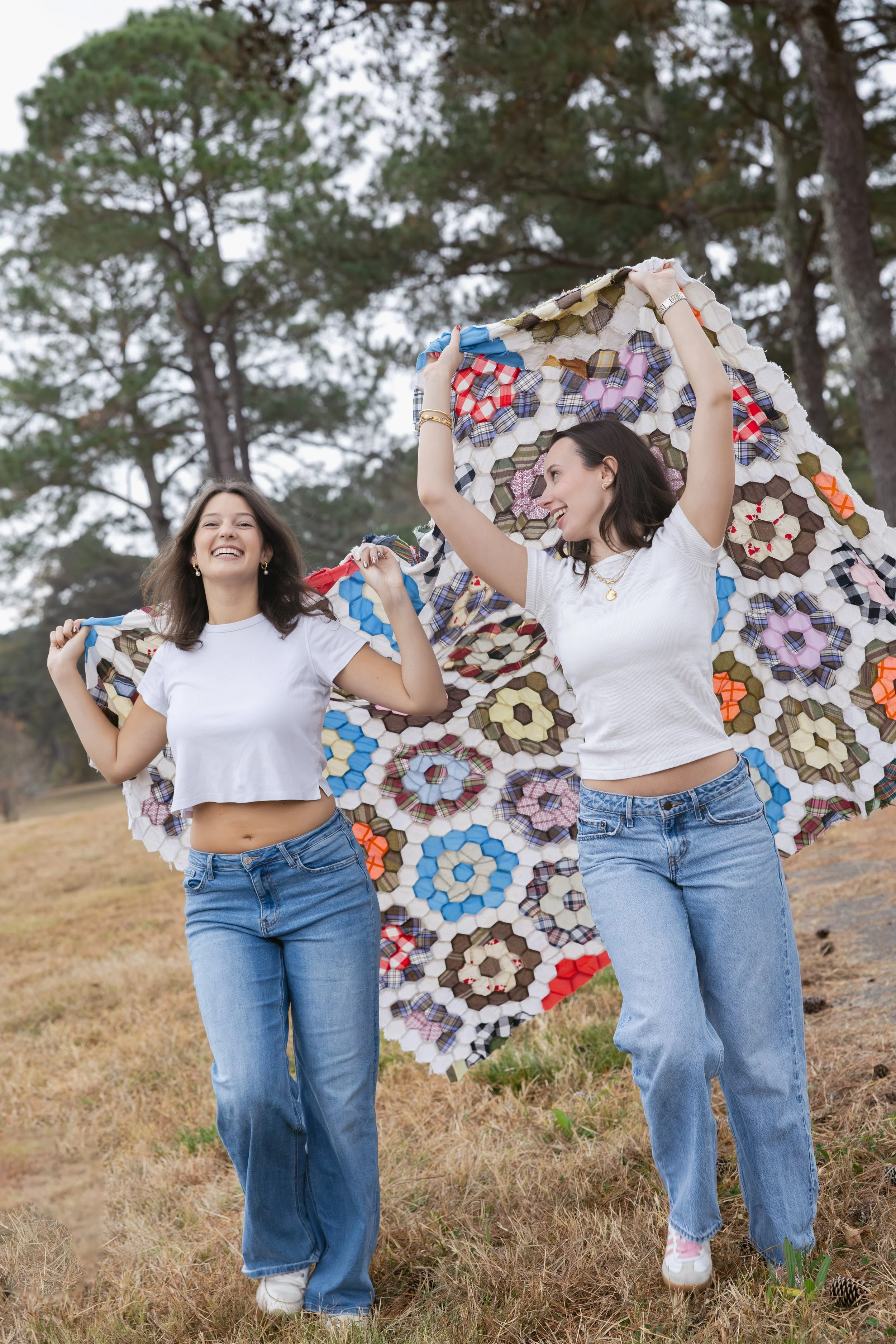 Two women smiling and holding a colorful quilt outdoors with trees in the background.