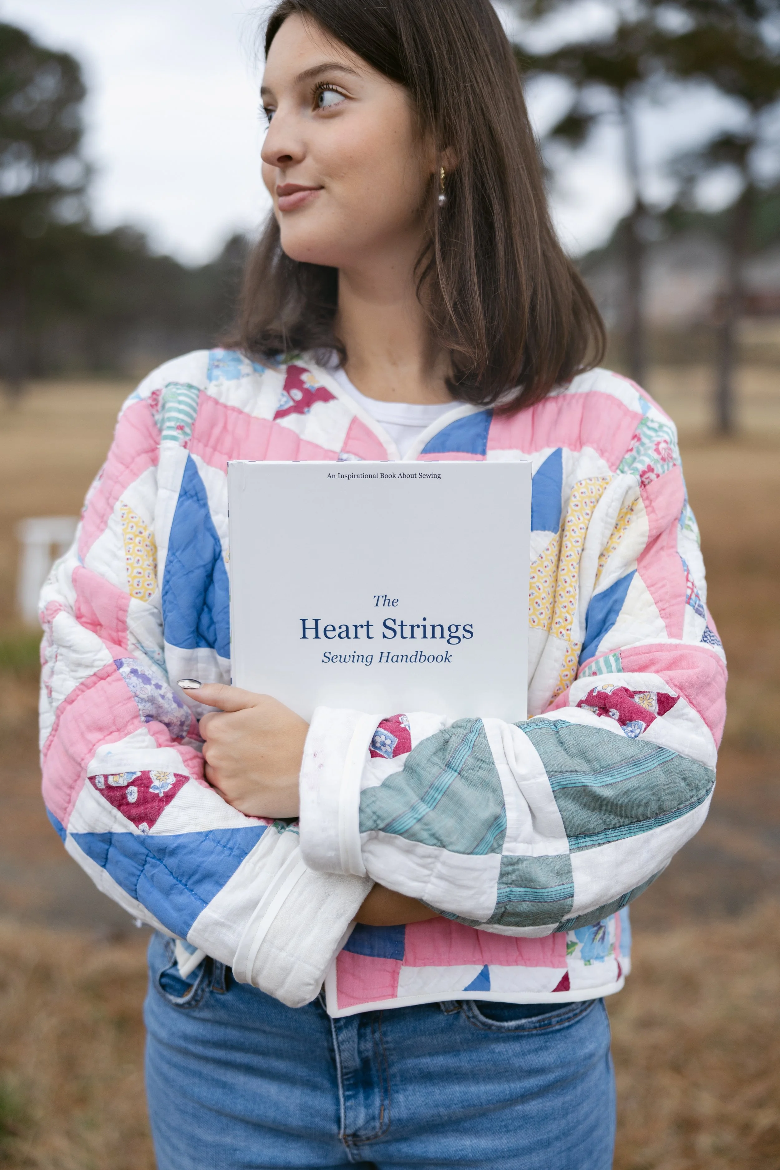 A young woman in a colorful patchwork quilted jacket holds a sewing book titled 'The Heart Strings Sewing Handbook' against an outdoor background with trees and a cloudy sky.