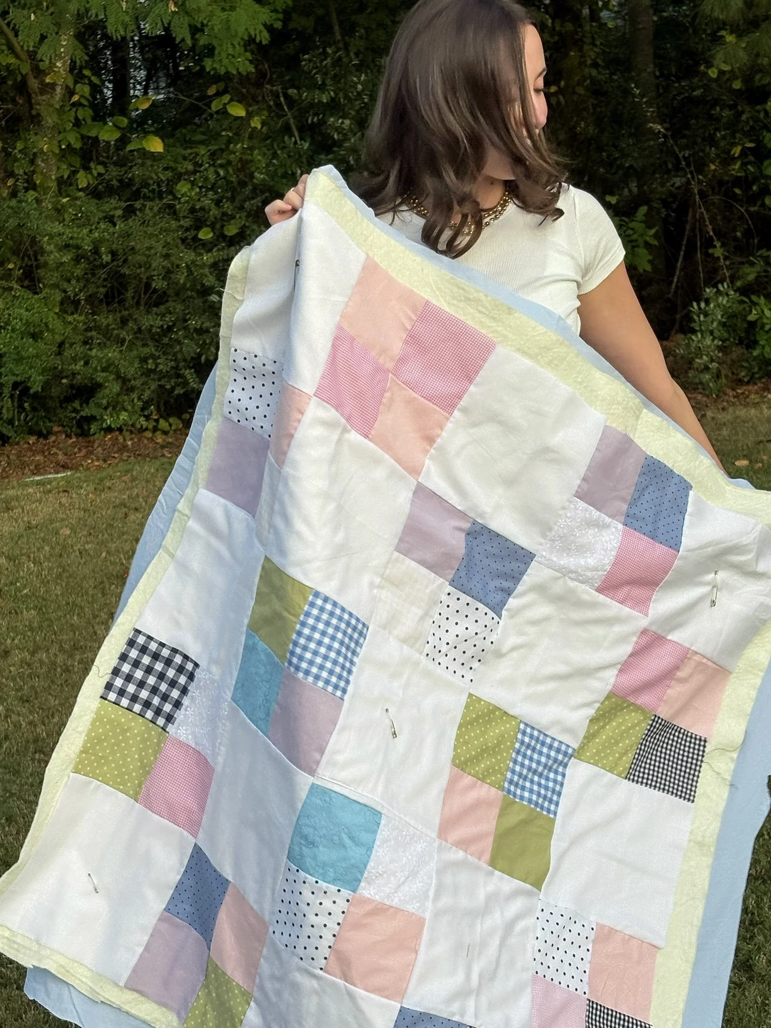 A woman holding a pastel-colored patchwork quilt outdoors with green trees in the background.