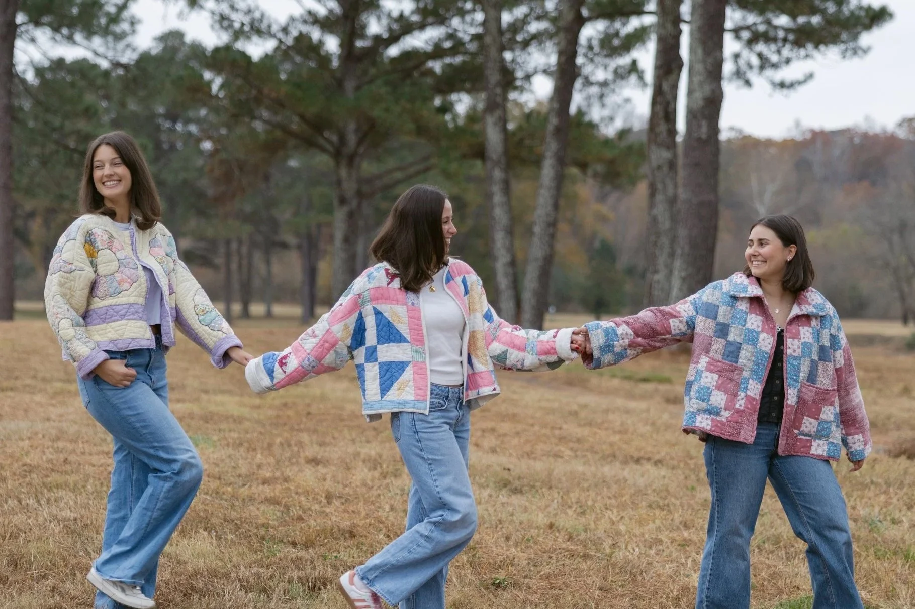 Three women holding hands and smiling in a park with trees, wearing colorful quilted jackets and jeans.
