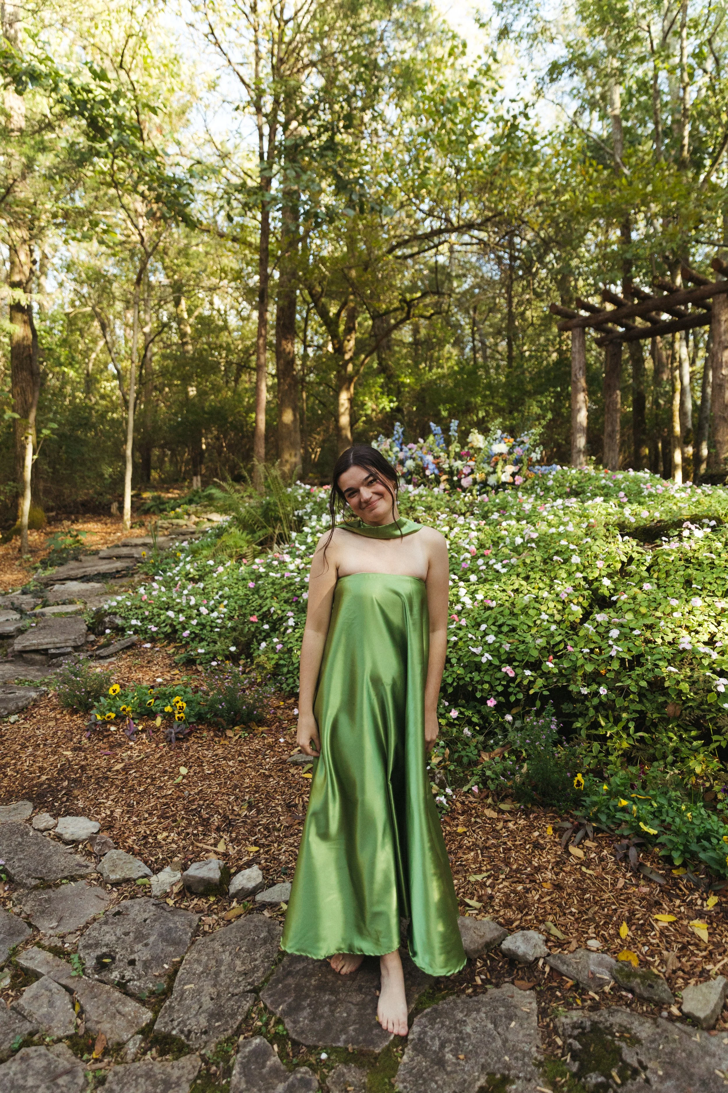 A young woman in a shiny green strapless dress poses barefoot on a stone pathway in a beautiful garden surrounded by lush green trees and blooming flowers.