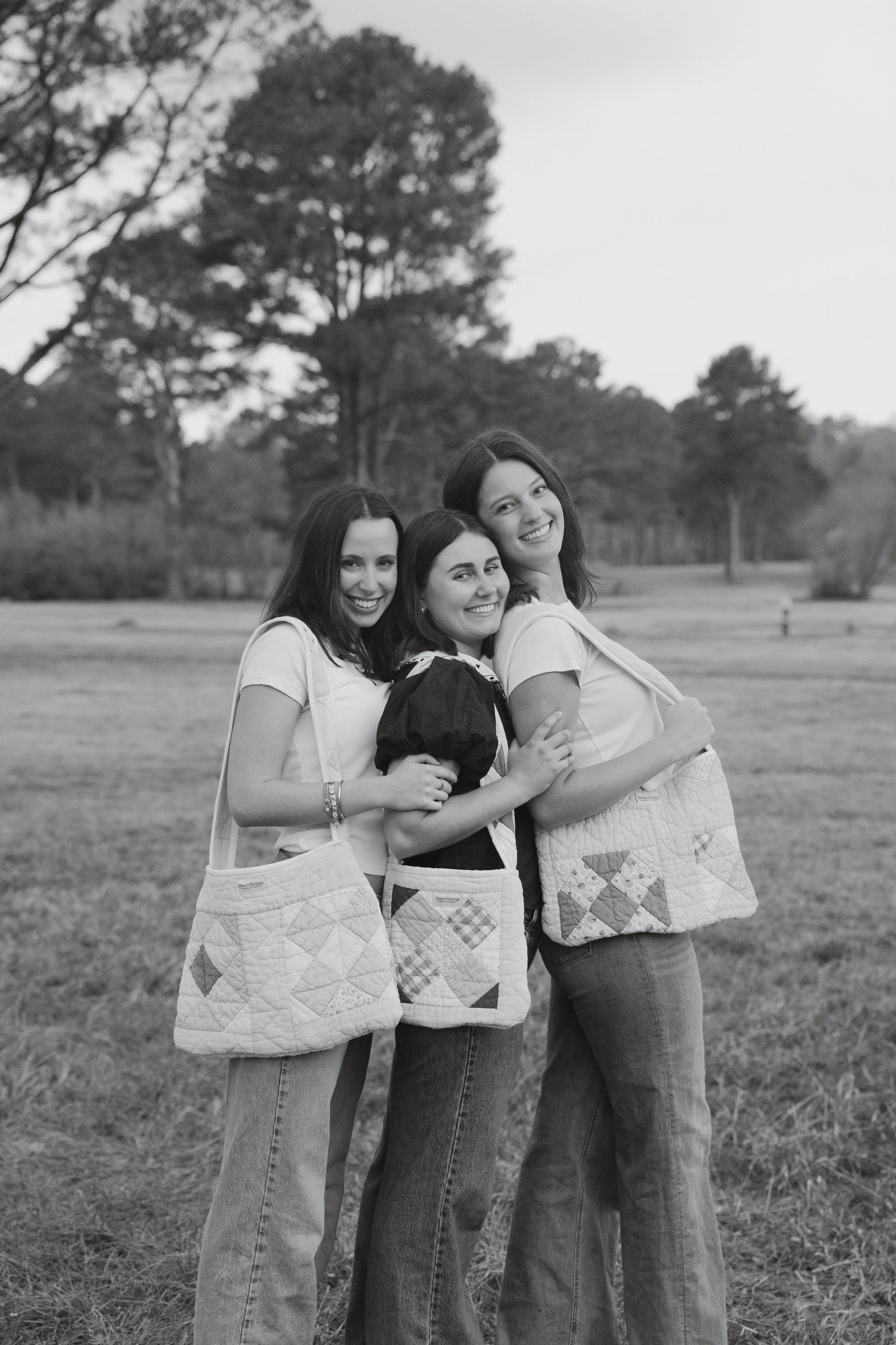 Black and white photo of three young women standing outdoors in a field, smiling and embracing each other, wearing casual clothes with quilted tote bags over their shoulders.