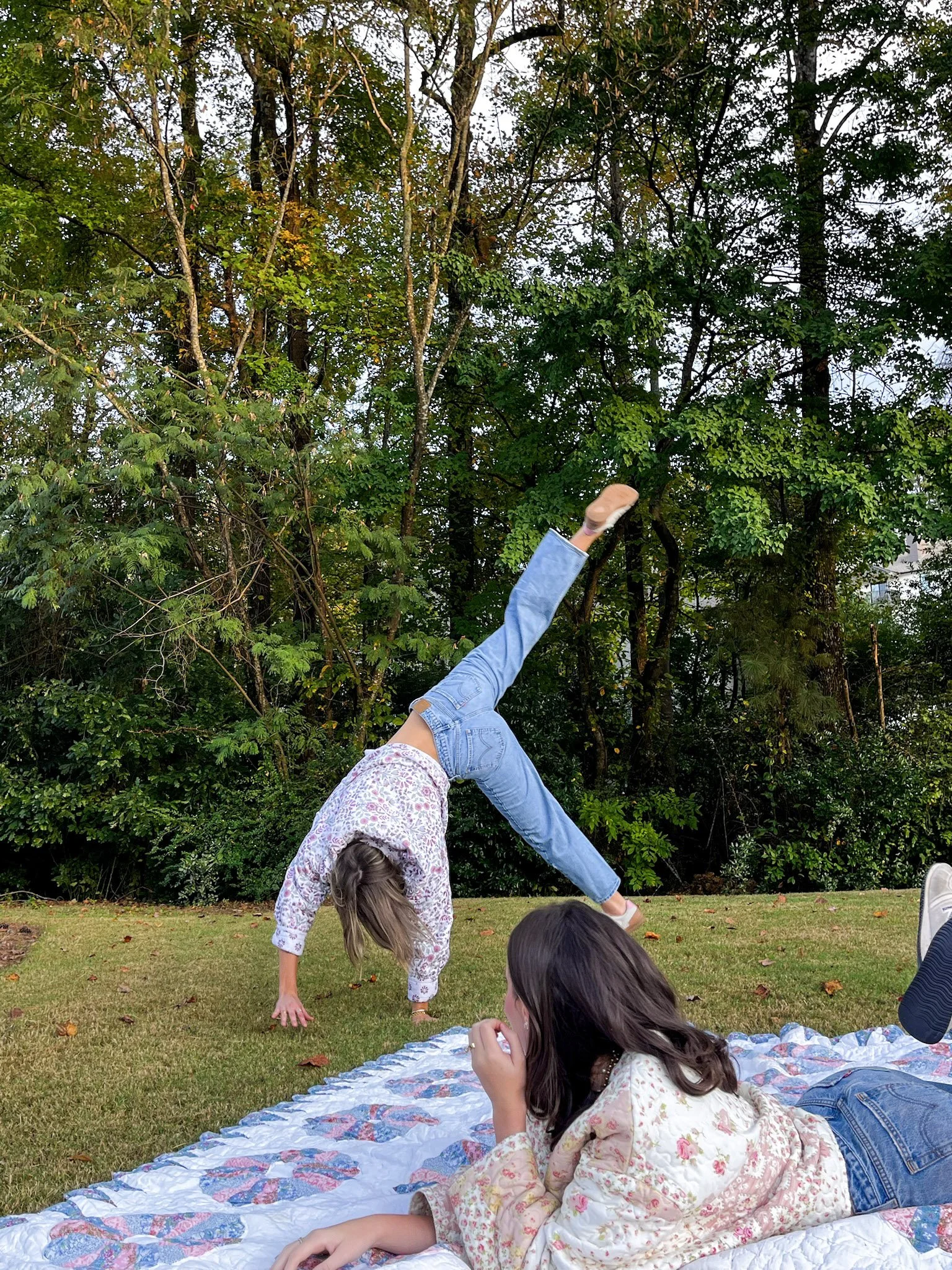 Two children, a girl doing a handstand and another lying on a blanket, in a backyard with trees.