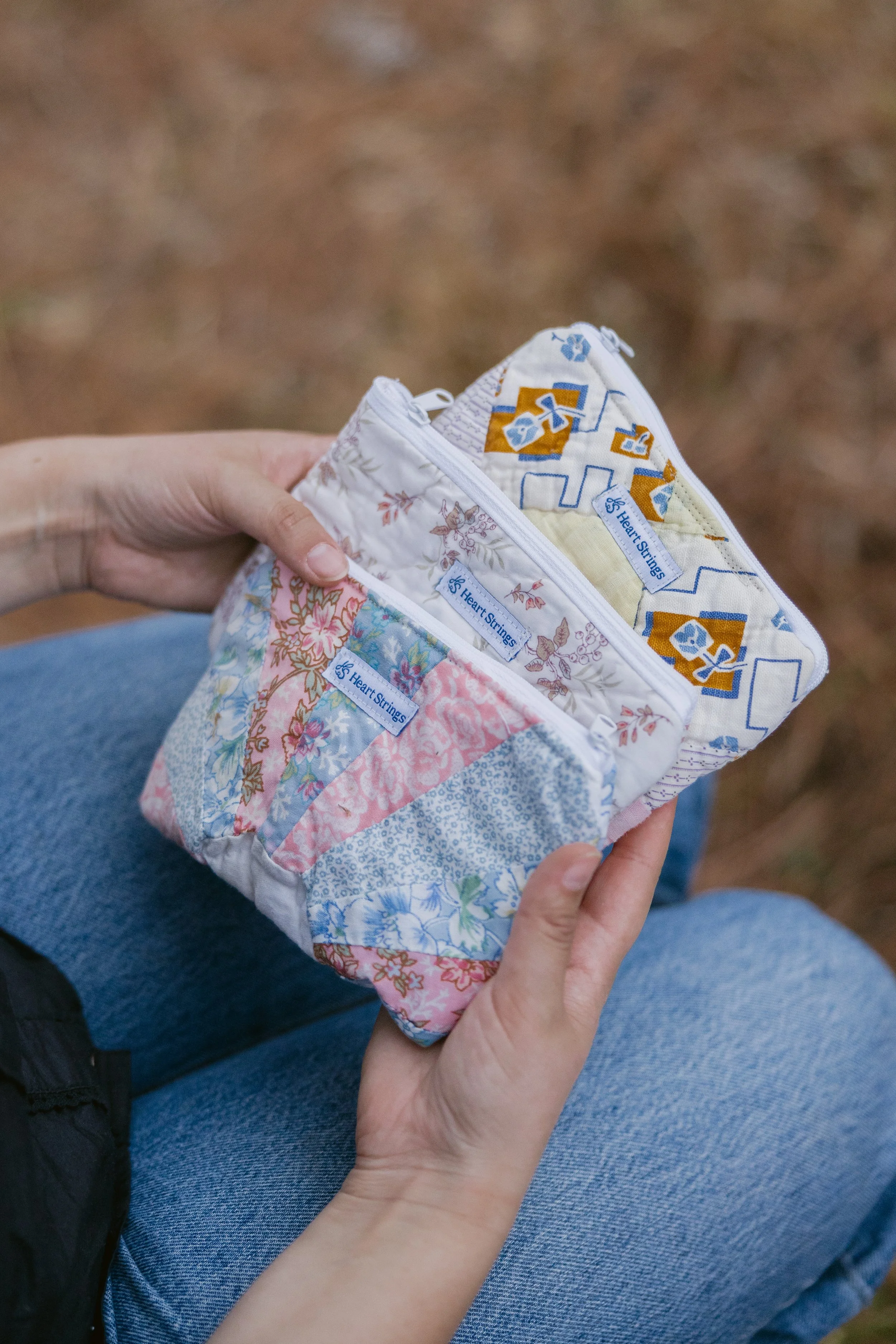 A person holding three small quilted fabric pouches with zippers, each with a blue label reading 'Heart Strings'. The person is sitting outdoors on a seat with their legs crossed, wearing blue jeans.
