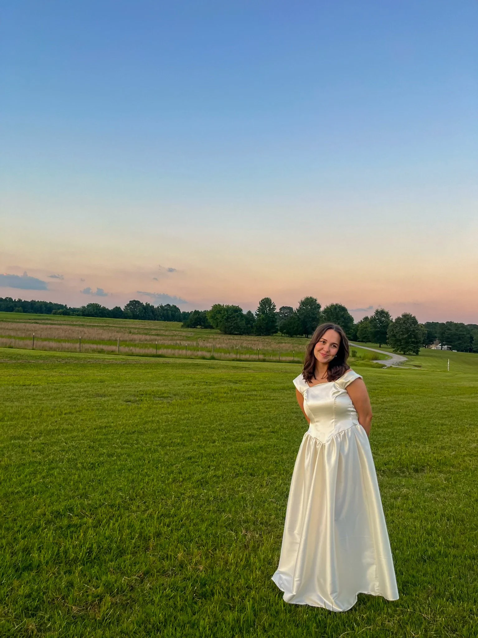Young woman in a white satin dress standing on a grassy field at sunset, with trees and a winding road in the background.
