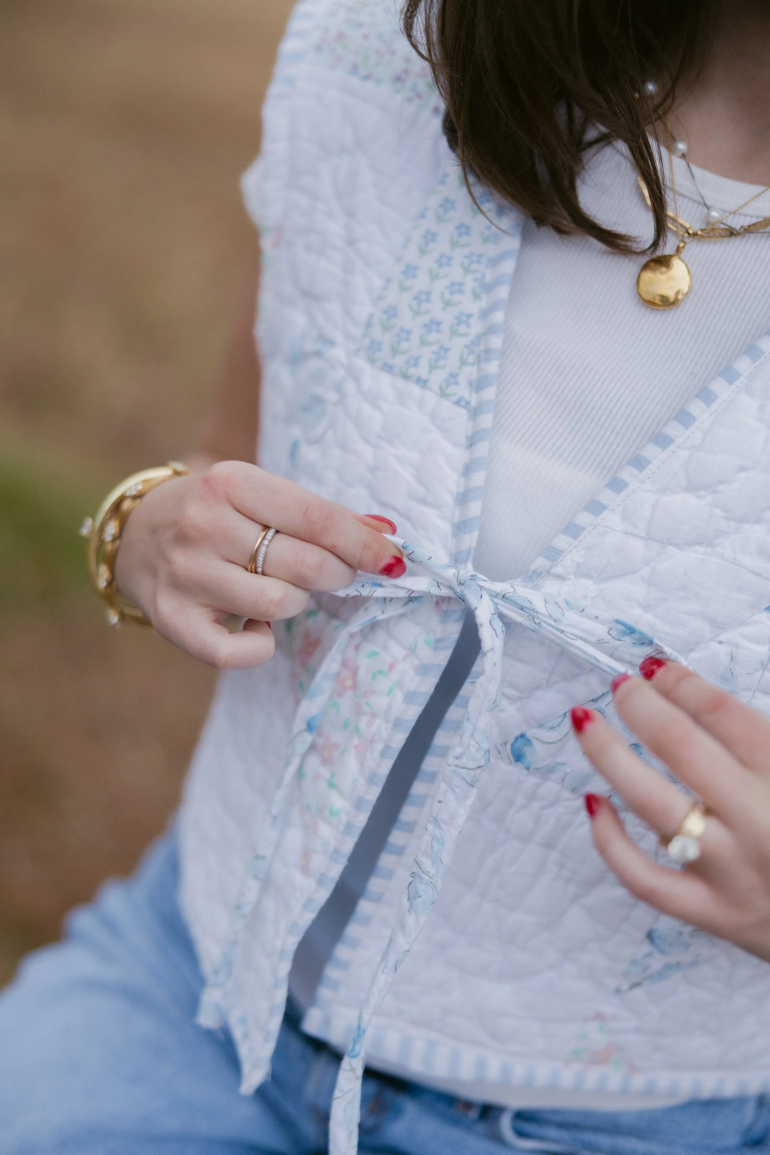 A woman tying the bow on a quilted vest, wearing jewelry and red nail polish.