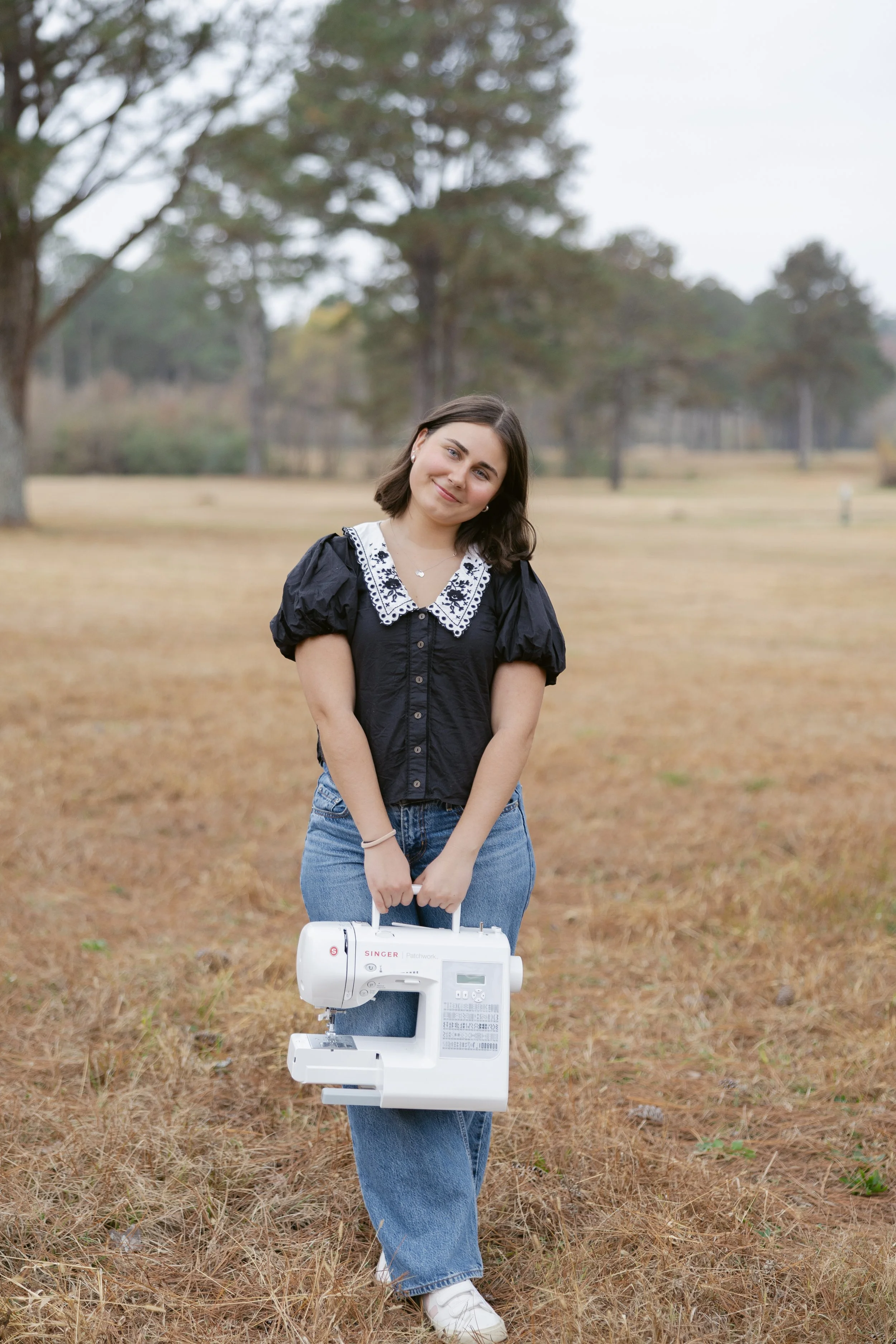 A young woman standing outdoors in a park with trees, holding a white sewing machine, wearing a black blouse with a decorative white collar and jeans.
