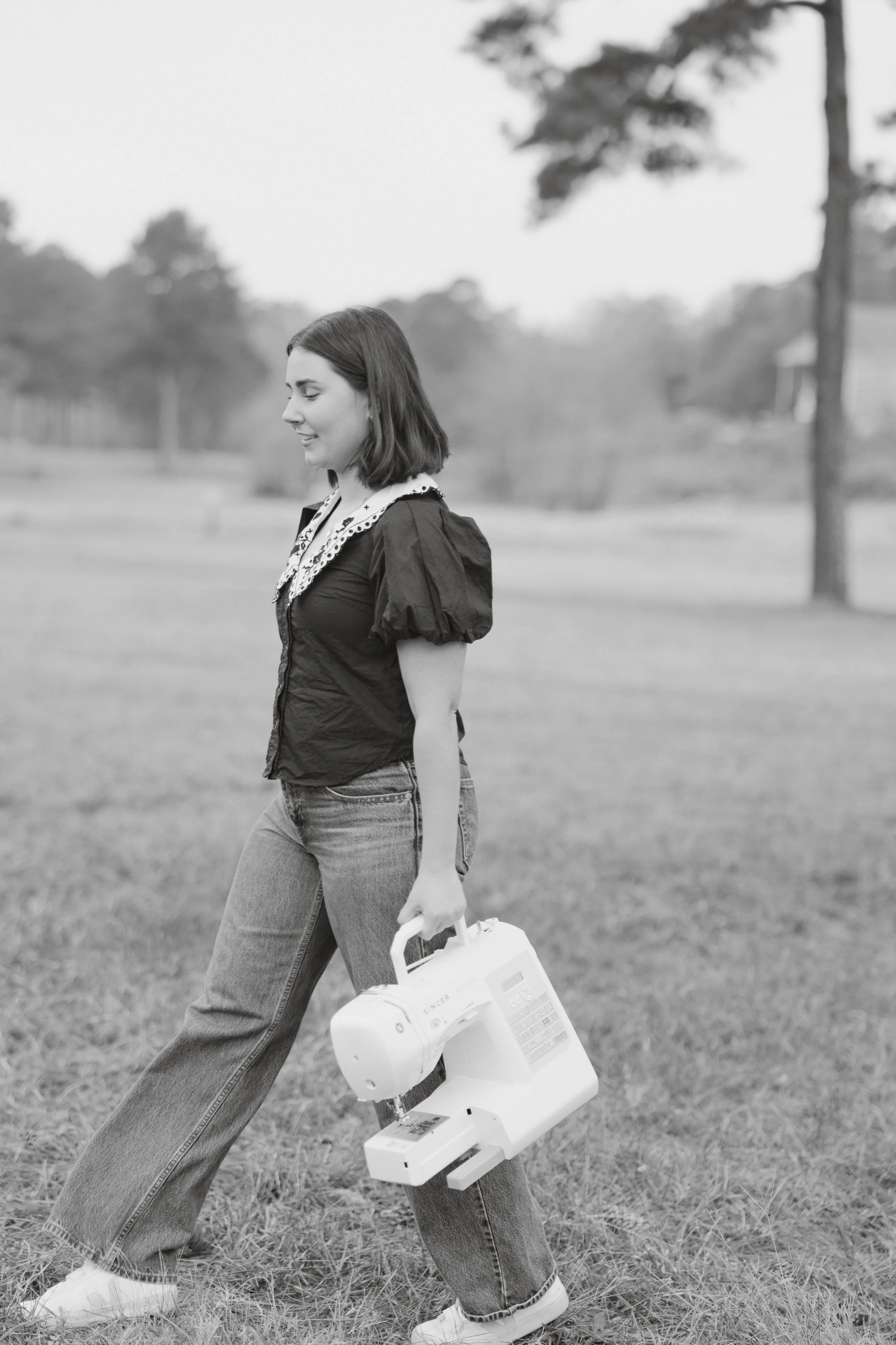 A young woman with shoulder-length hair walking outdoors on grass, carrying a sewing machine, and smiling while looking down.