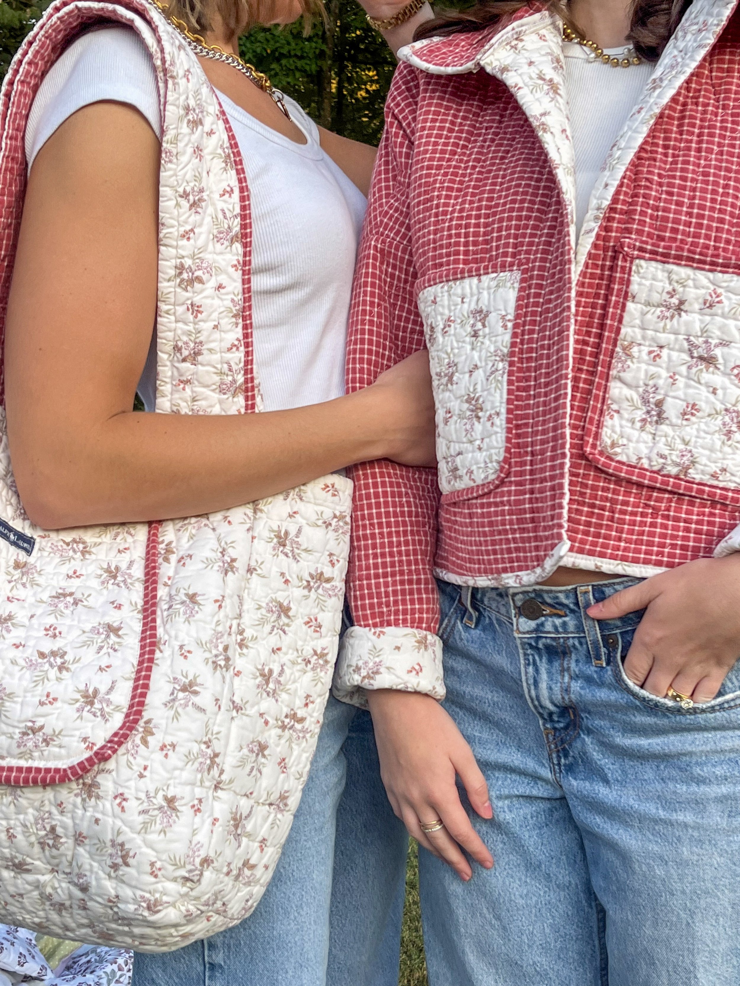 Close-up of two women standing close together, wearing vintage-style vests and denim jeans. One woman is holding the other's arm, and they are outdoors with greenery in the background.