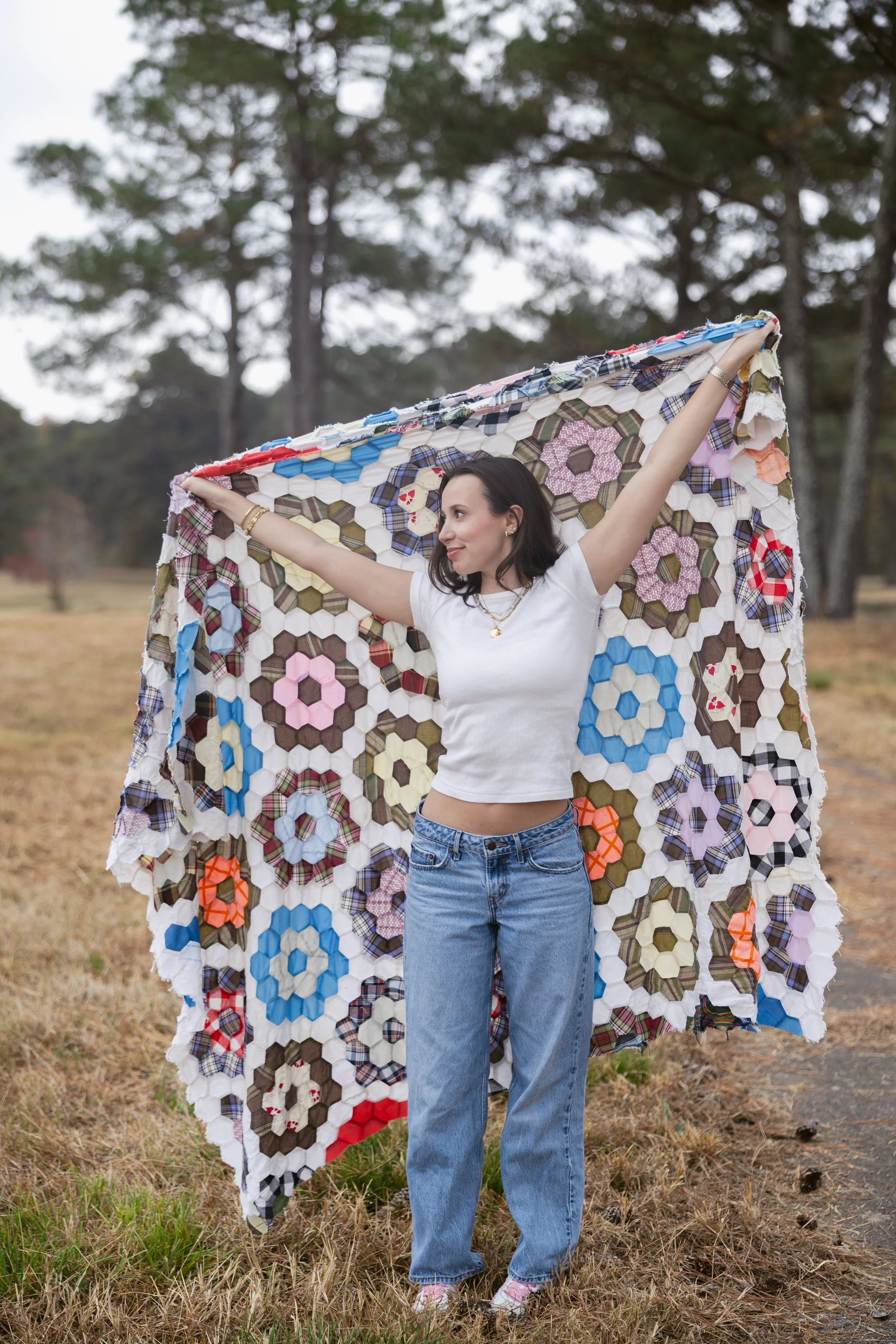 A woman holding a quilt outdoors in a grassy area with trees in the background.