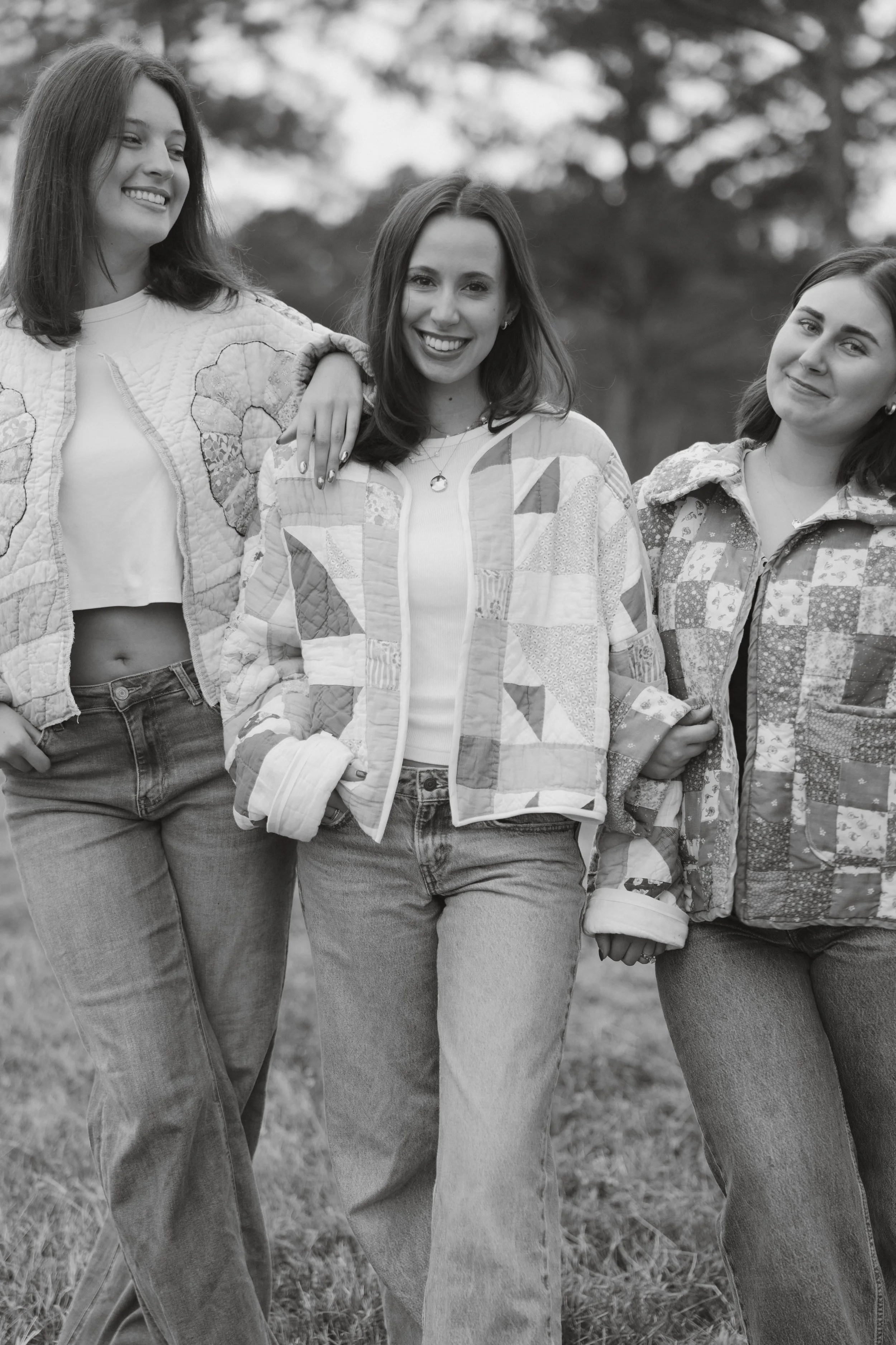 Three women standing outdoors, smiling, wearing casual jackets and jeans, in a natural setting with trees in the background.