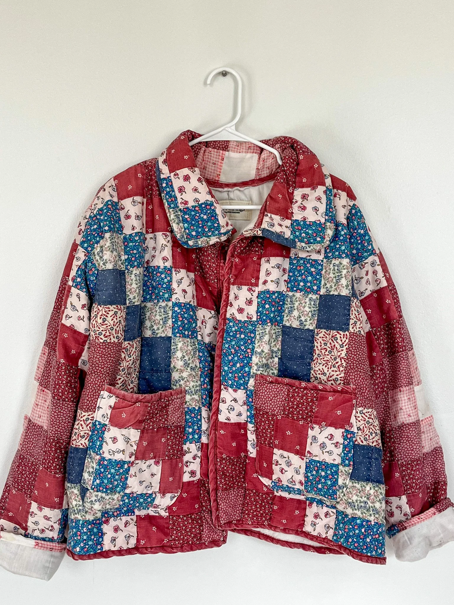 Patchwork quilted jacket with red, white, and blue fabric squares, hanging on a white wall.