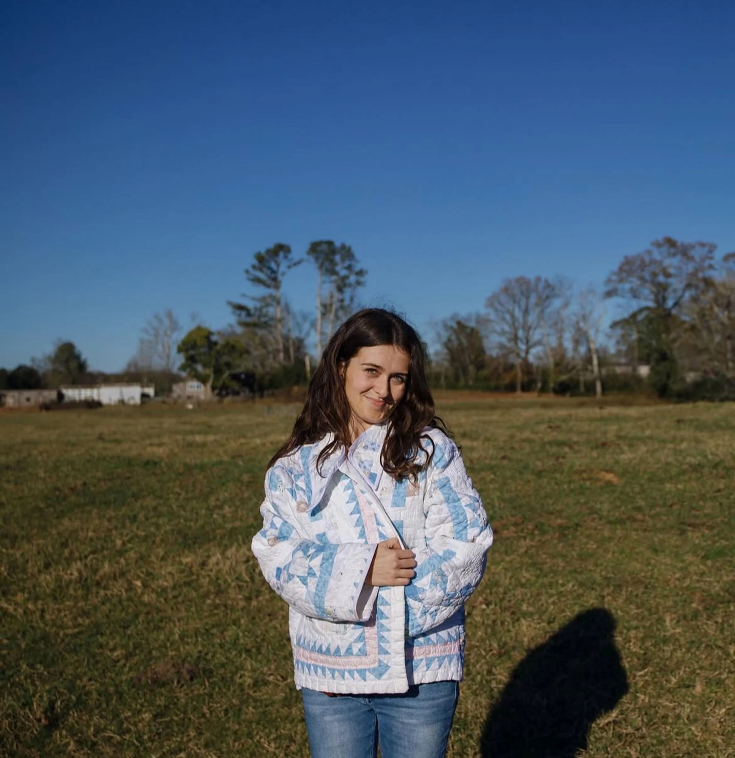 Young woman with long brown hair standing outside in a field, wearing a patterned white and blue quilted jacket and jeans, smiling at the camera with trees and a clear blue sky in the background.