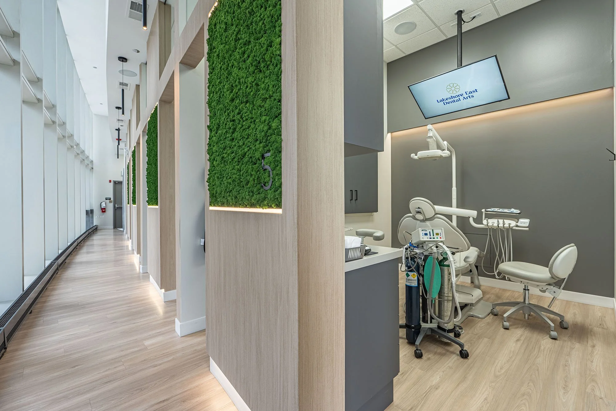 A modern dental clinic interior with a hallway on the left and a dental treatment room on the right. The hallway has wood flooring, white walls, and green moss wall decor with the number 5. The dental room has gray walls, a dental chair, and equipment, with a large monitor displaying 'Lakeshore East Dental Arts.'