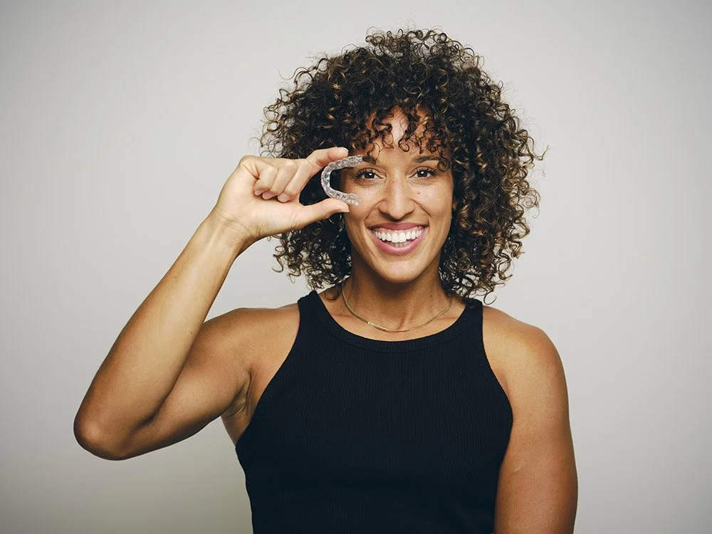 Smiling woman with curly hair holding a clear dental retainer in front of her eye.