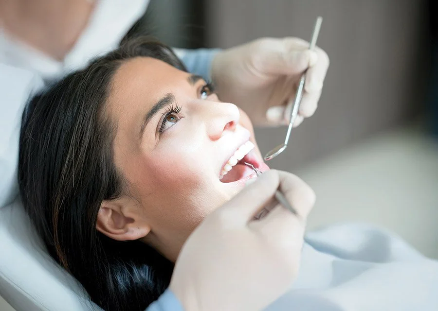 A woman lying back in a dental chair with her mouth open, being examined by a dentist using dental tools.