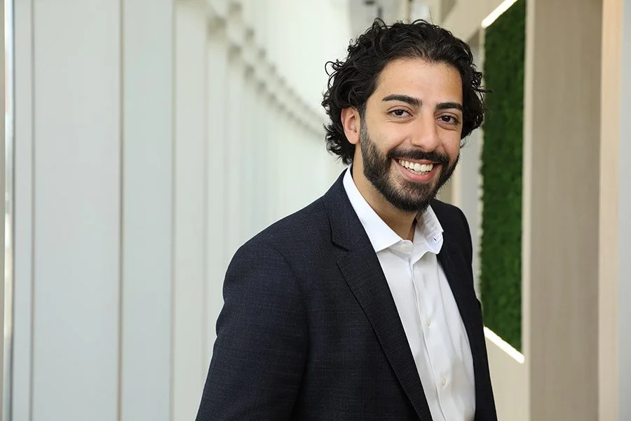 Smiling man with dark curly hair and beard in formal suit standing in modern office hallway.