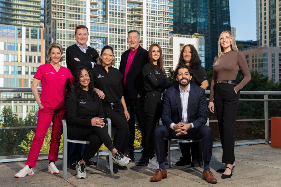 Group of nine diverse professionals, including healthcare workers in scrubs and business people in suits, posing outdoors on a city balcony with tall buildings in the background.