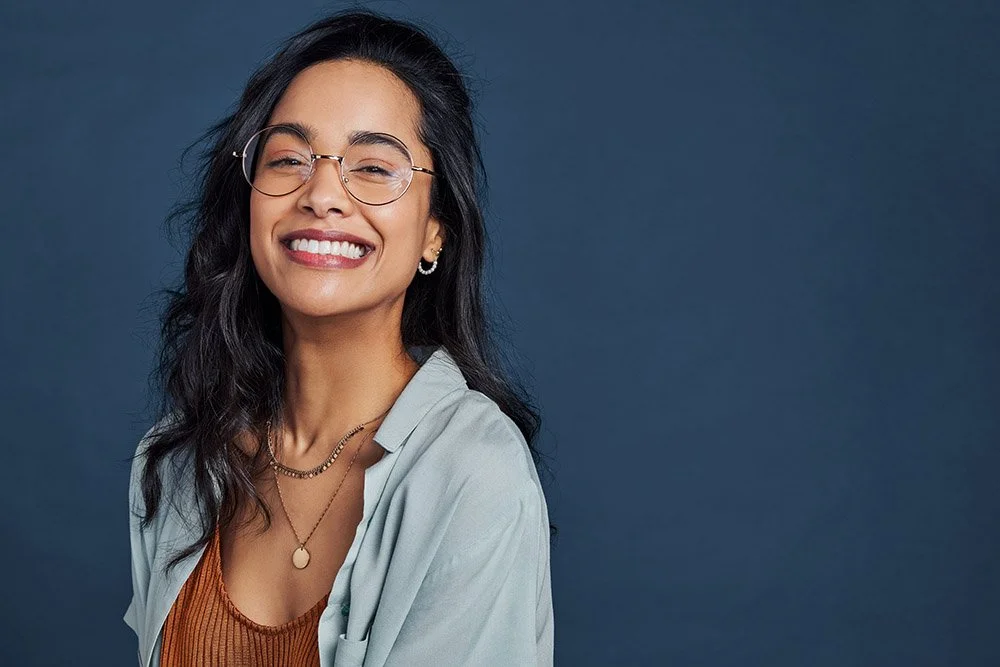 A smiling woman with long dark hair, glasses, and hoop earrings, wearing a light-colored shirt over an orange top, against a dark blue background.