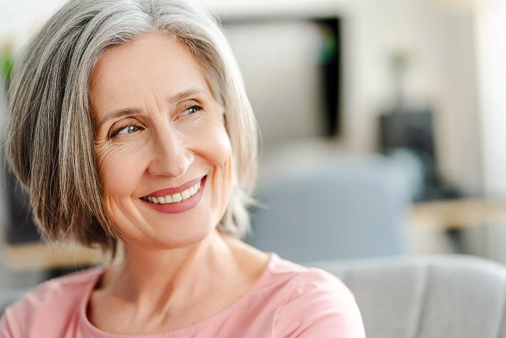 Smiling middle-aged woman with gray hair in an indoor setting, possibly an office or living room.