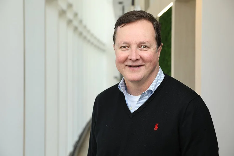A man with short dark hair, wearing a light blue collared shirt and a black sweater with a small red polo logo, standing in a bright corridor with white walls and a green background.