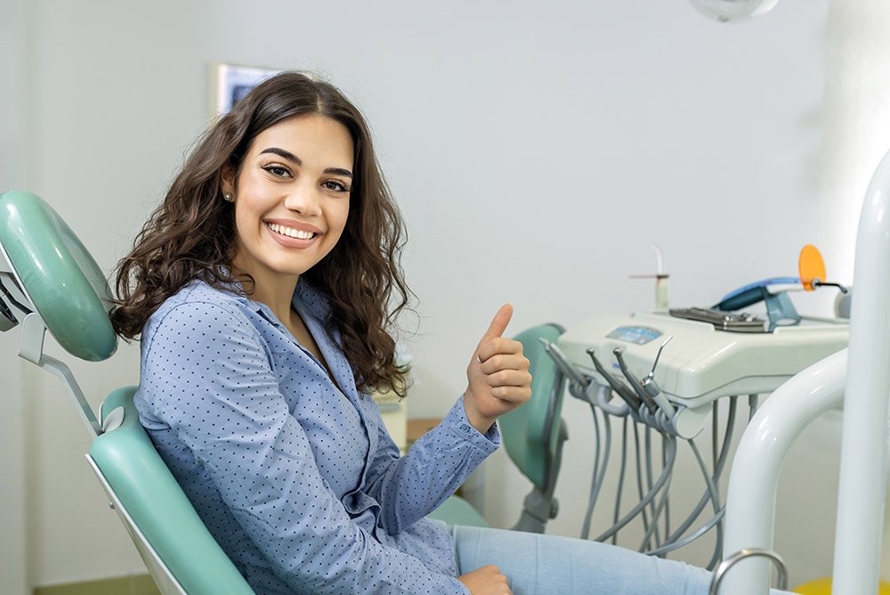 A woman sitting in a dental chair at a dentist's office, giving a thumbs up and smiling.