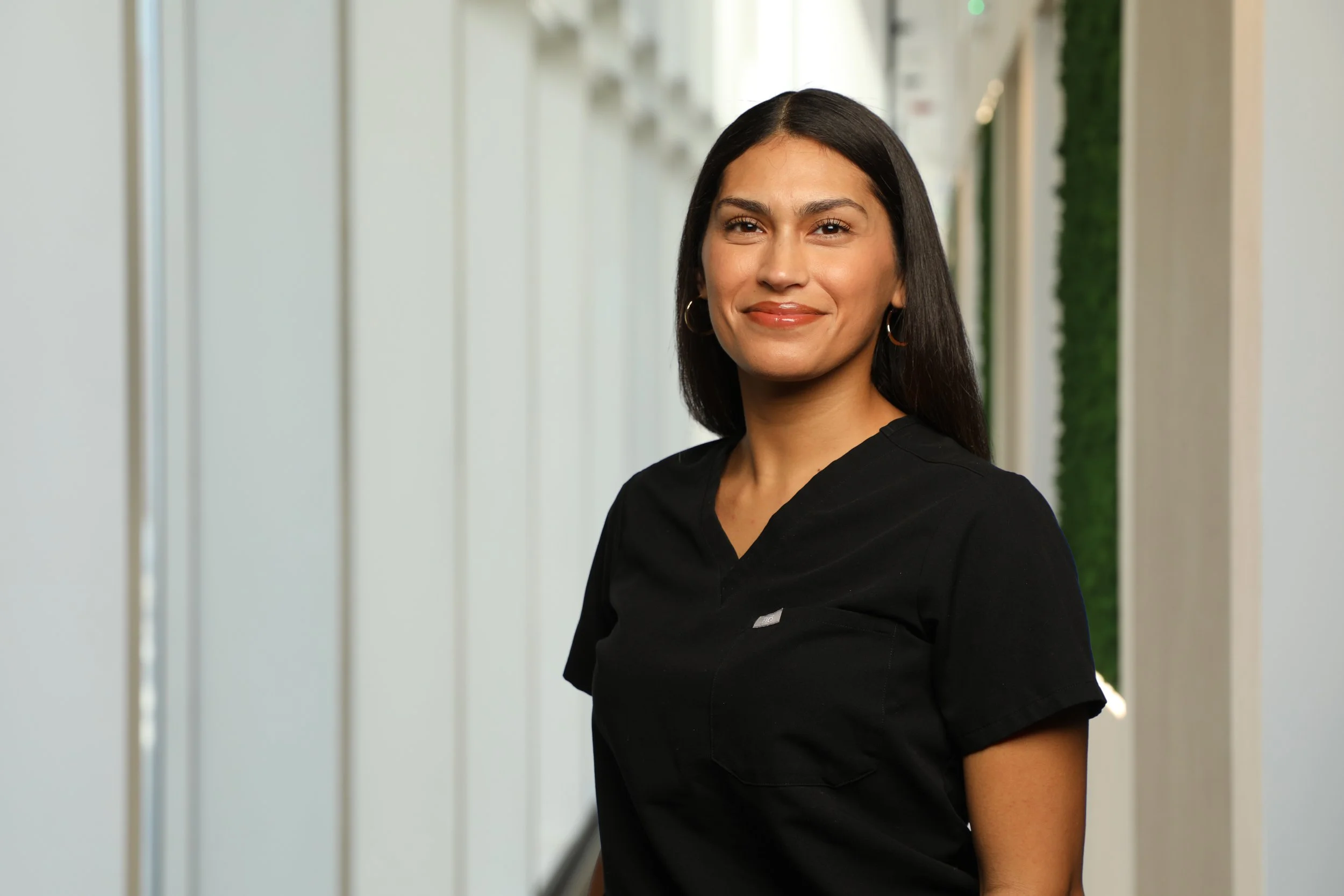 A woman with dark hair, wearing a black top, smiling in a modern indoor setting with light-colored walls and green accents.