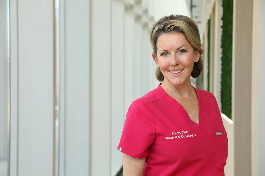 A smiling woman in a pink medical uniform standing in a bright corridor.