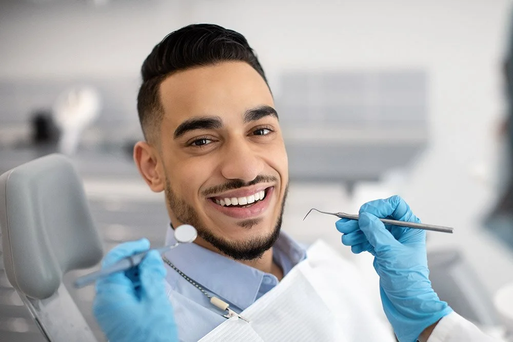 A young man at the dentist smiling while getting a dental checkup, wearing gloves and a dental bib.