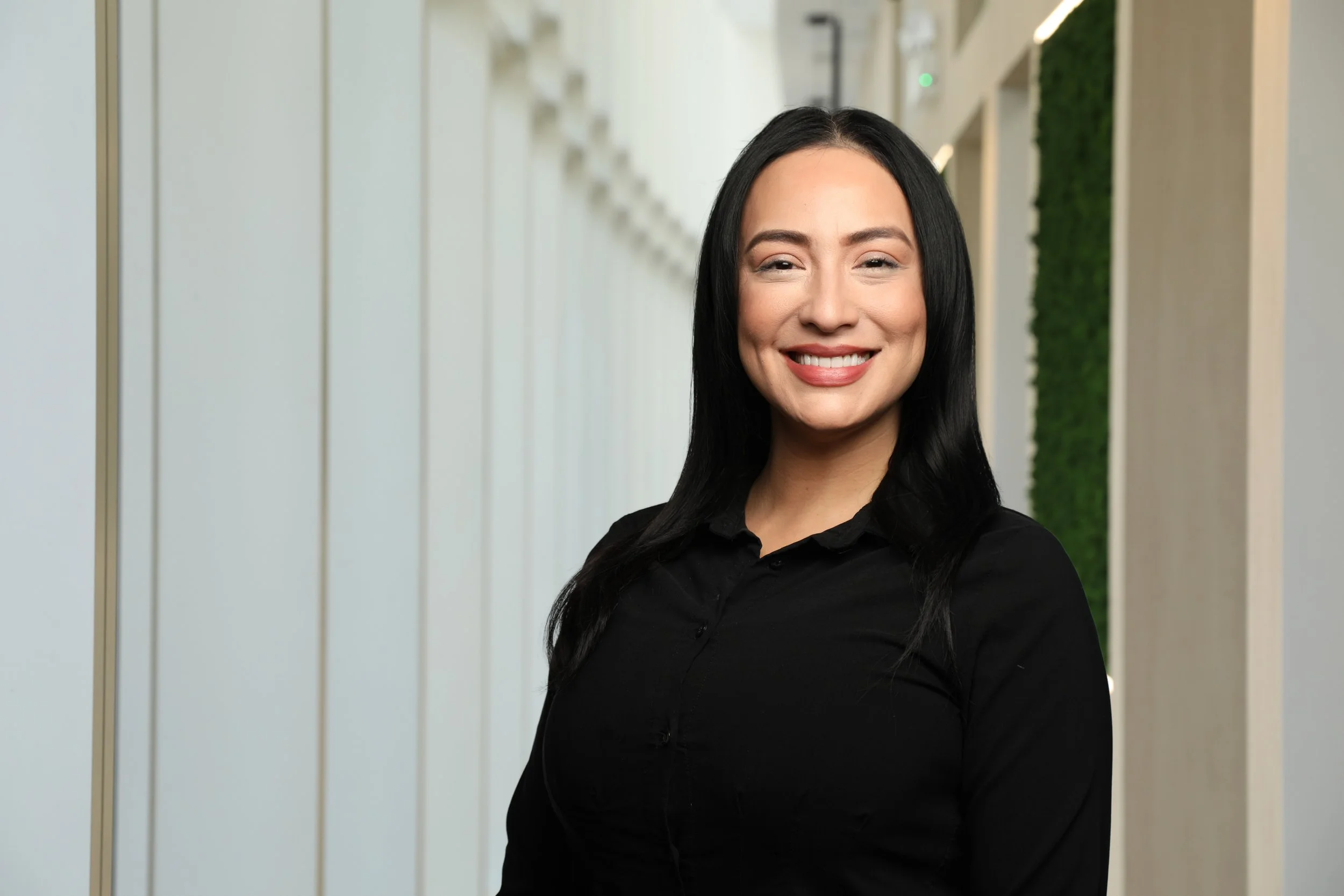 A woman with long black hair smiling at the camera, wearing a black shirt, standing in an indoor corridor with light-colored walls and a green wall in the background.