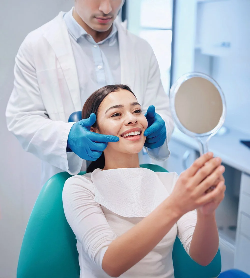 A woman sitting in a dental chair holding a mirror, smiling, with a dentist examining her teeth and holding her cheeks, in a dental clinic.