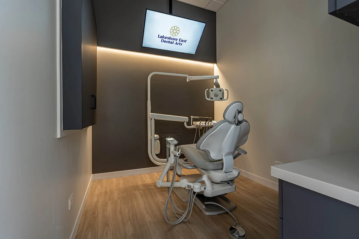 Dental exam chair in a modern dental clinic with a wall-mounted monitor displaying 'Lakeshore East Dental Arts' logo.