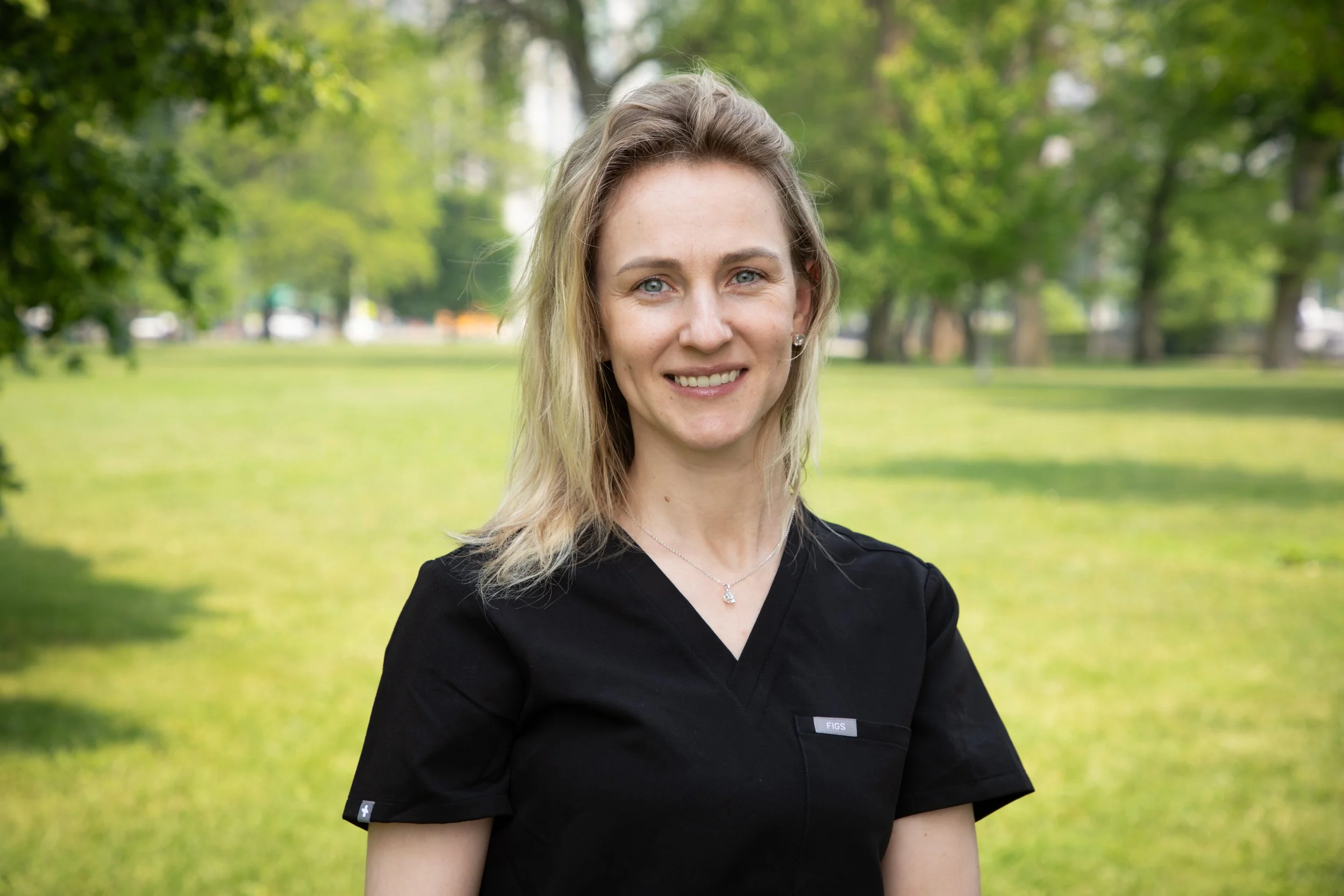 A woman with blonde hair and blue eyes smiling outside, wearing a black shirt and a silver necklace, with green trees and grass in the background.