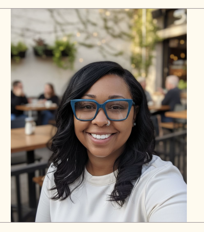 A woman with black hair, wearing blue glasses and a white shirt, smiling at a social outdoor setting with other people in the background.