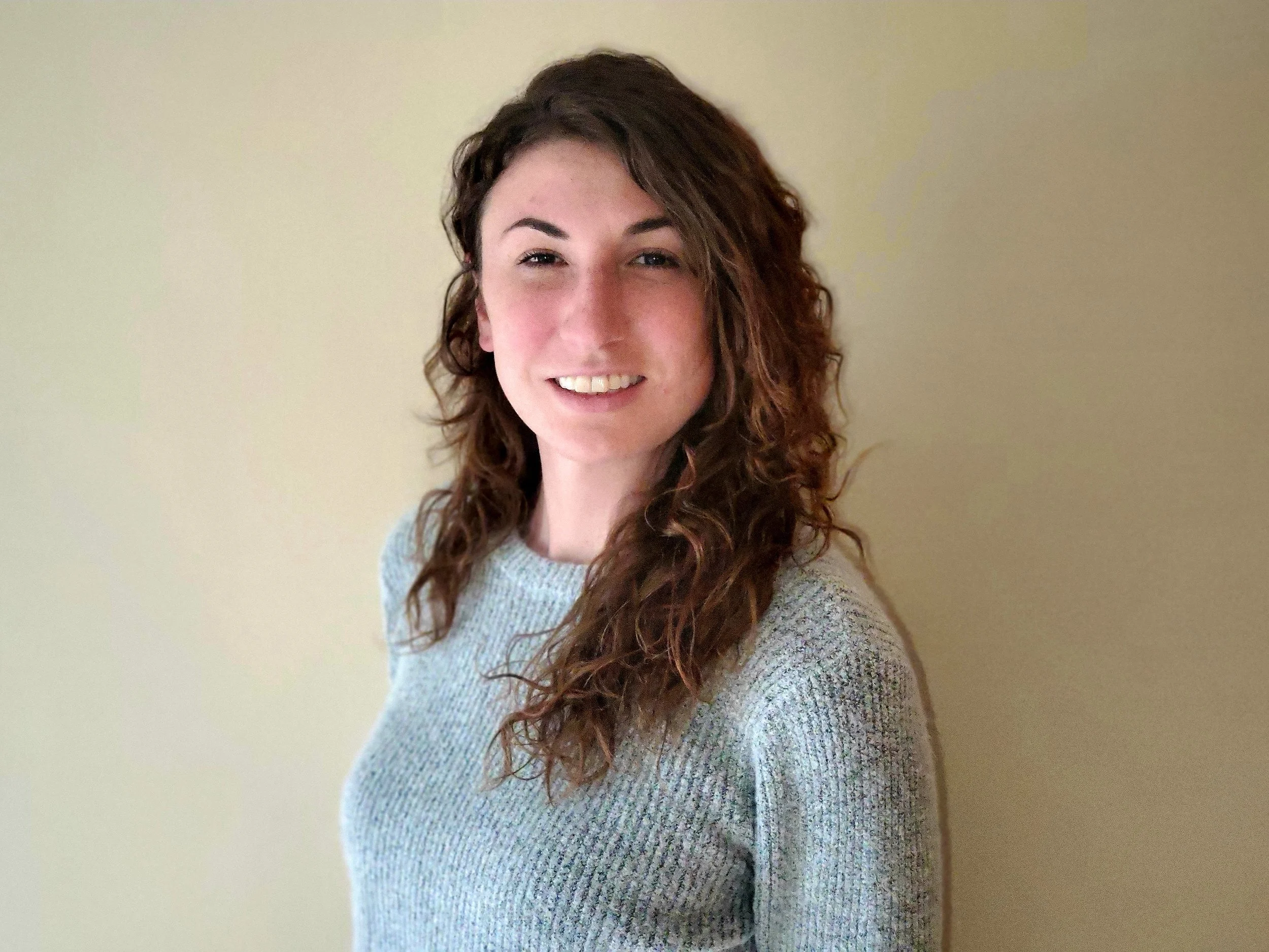 A young woman with long, curly brown hair smiling at the camera, wearing a gray sweater, standing against a plain beige wall.