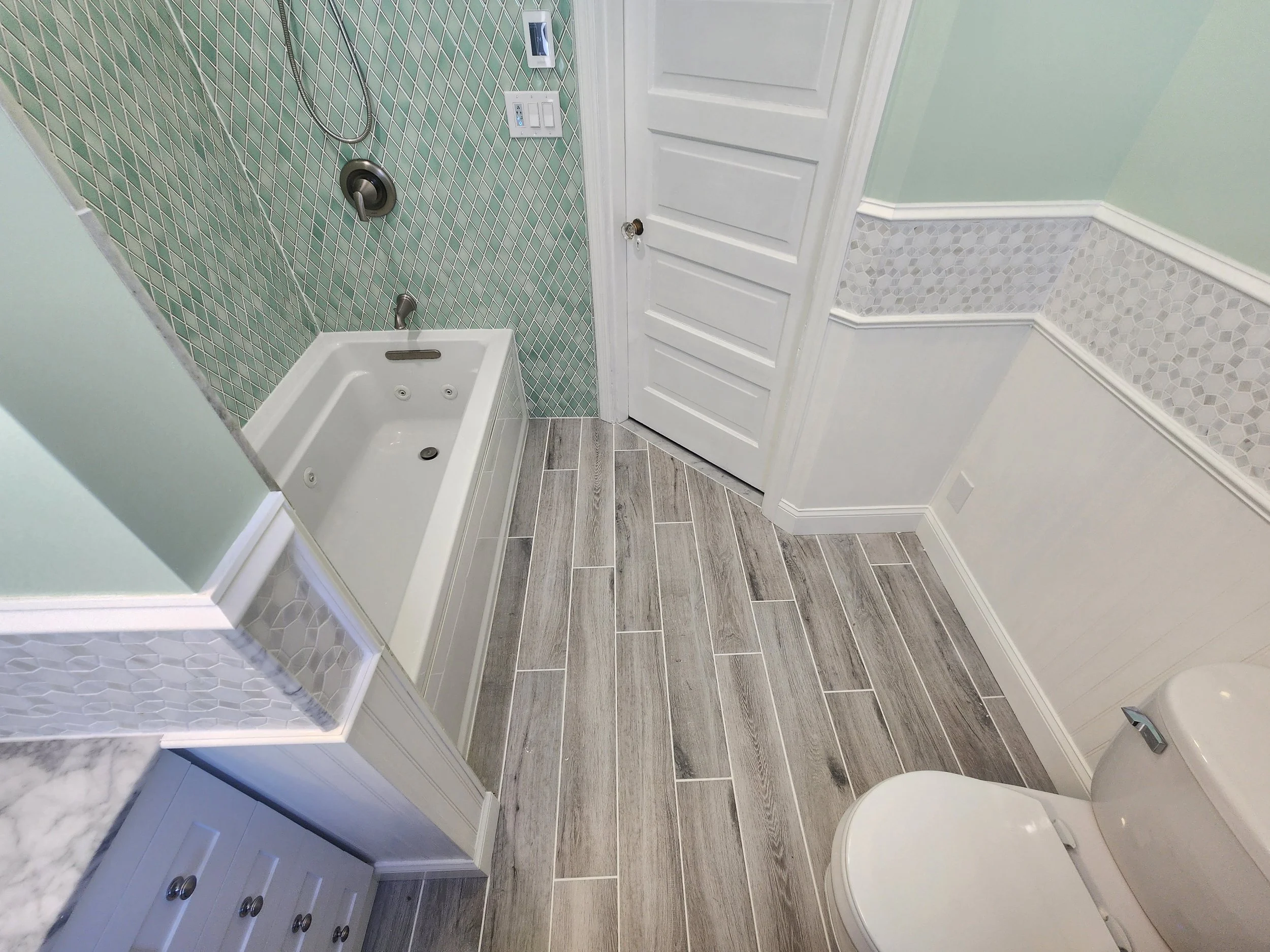 Bathroom with wood-look tile flooring, a white toilet, vanity with marble countertop, shower area with green tile walls and glass partition, and a white door.