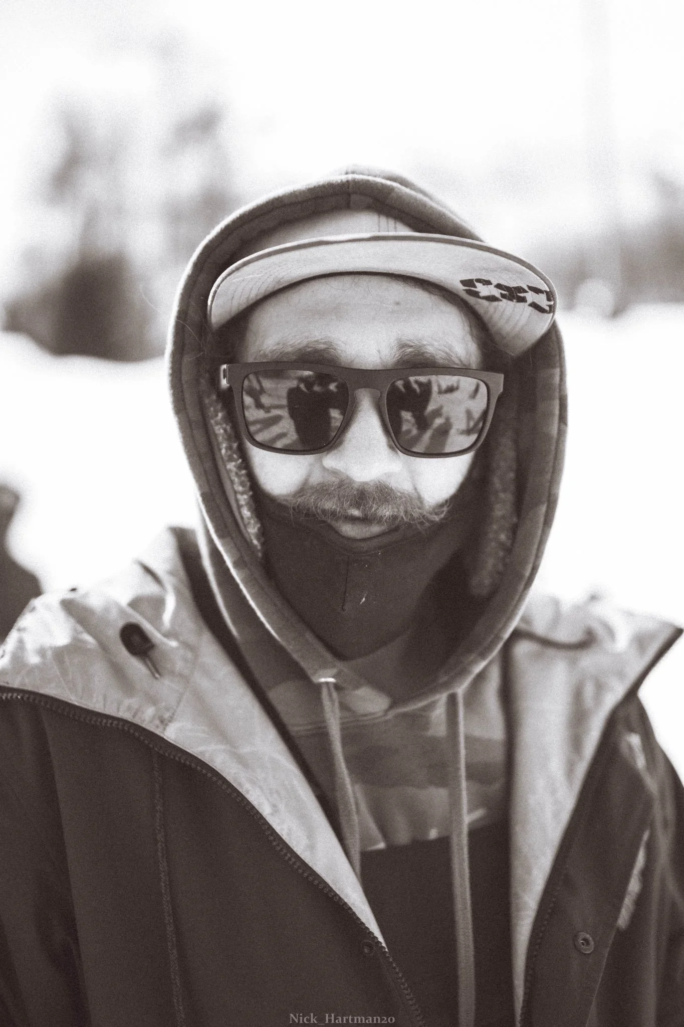 A person wearing sunglasses, a hoodie, a cap, and a face covering outdoors in a snowy environment.