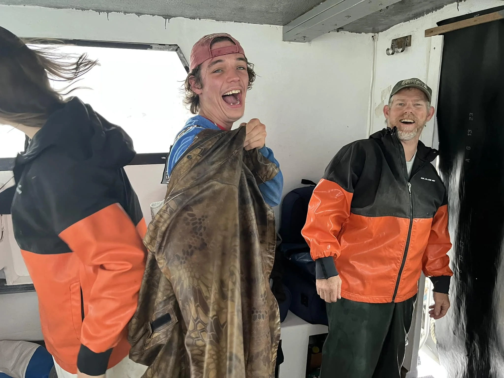 Three people on a boat, wearing rain jackets. One person in a red cap smiles, holding a patterned jacket. Another person in an orange and black jacket looks to the left, while the third, also in orange and black, with a cap, smiles at the camera.